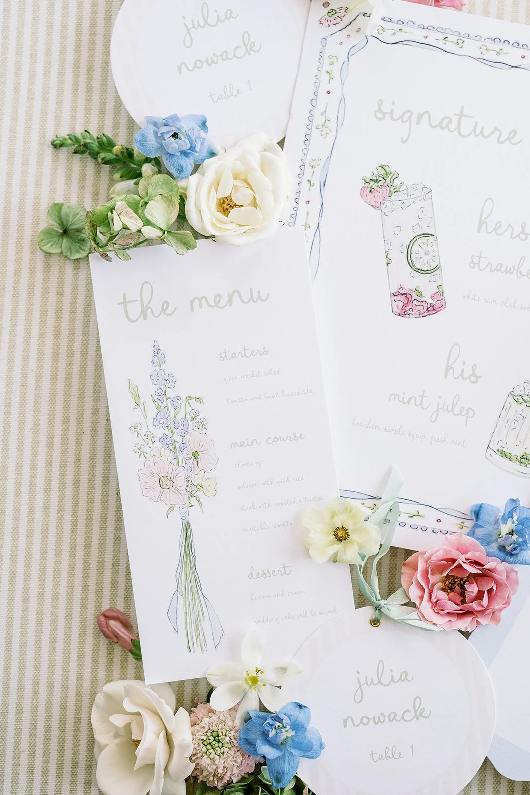 Wedding day-of paper design with hand-drawn style menus, floral decorations, and place cards on a striped tablecloth.