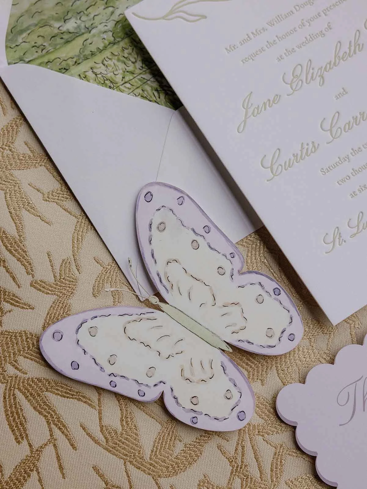 Gold patterned tablecloth with invitations and a butterfly cut-out over top of it for whimsical wedding stationery