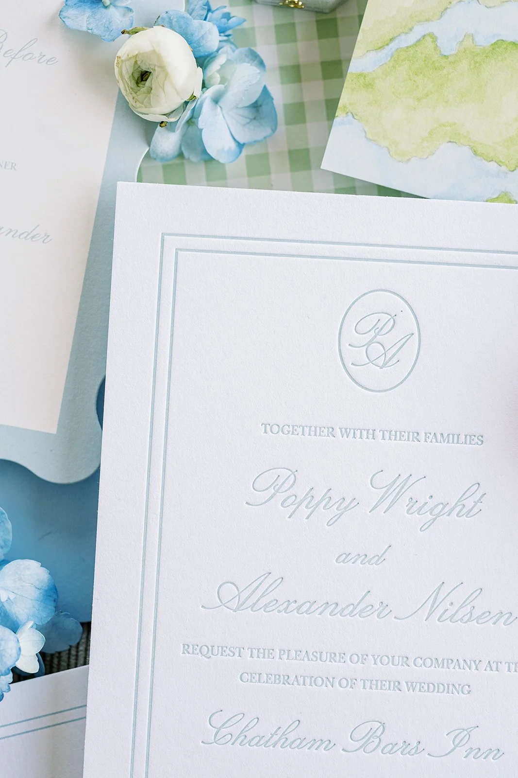 Custom wedding monogram on a wedding invitation on top of various coastal-themed wedding stationery and blue and white florals