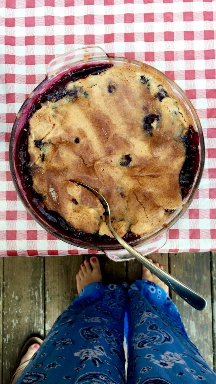 Overhead view of a blueberry cobbler in a baking dish on a red and white checkered tablecloth, with a spoon resting in it, and a person standing next to it.