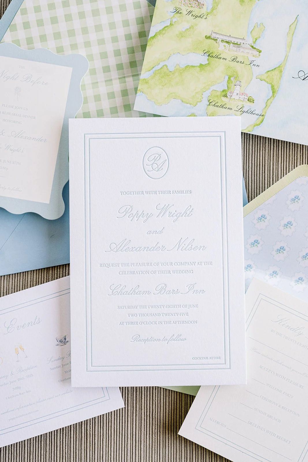 Wedding invitation with elegant script text, monogram at top, surrounded by other wedding stationery and a colorful watercolor map of a coastal area.
