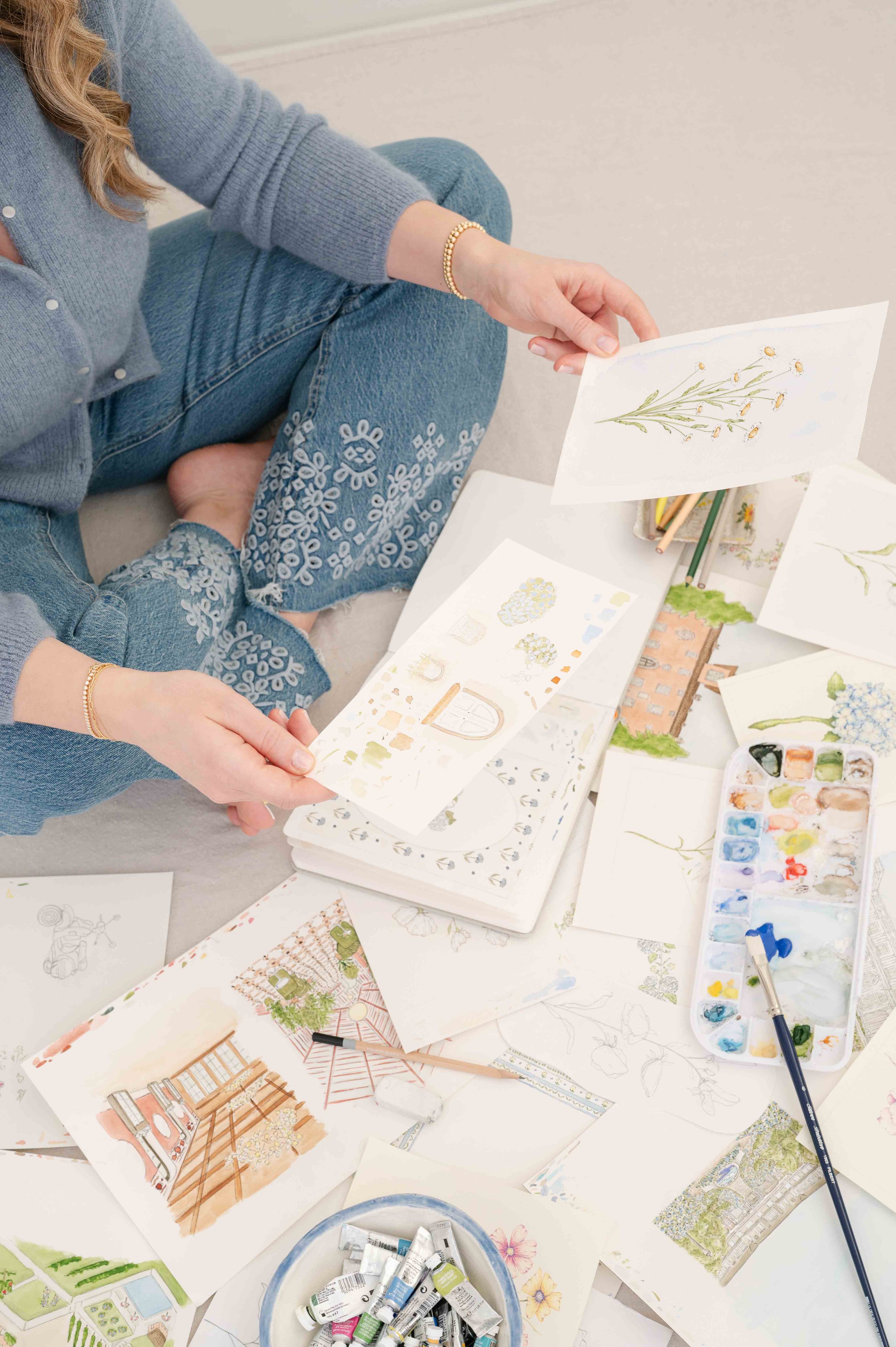Person sitting on the floor, surrounded by watercolor paintings, paint tubes, brushes, and paper, working on wedding venue illustrations.