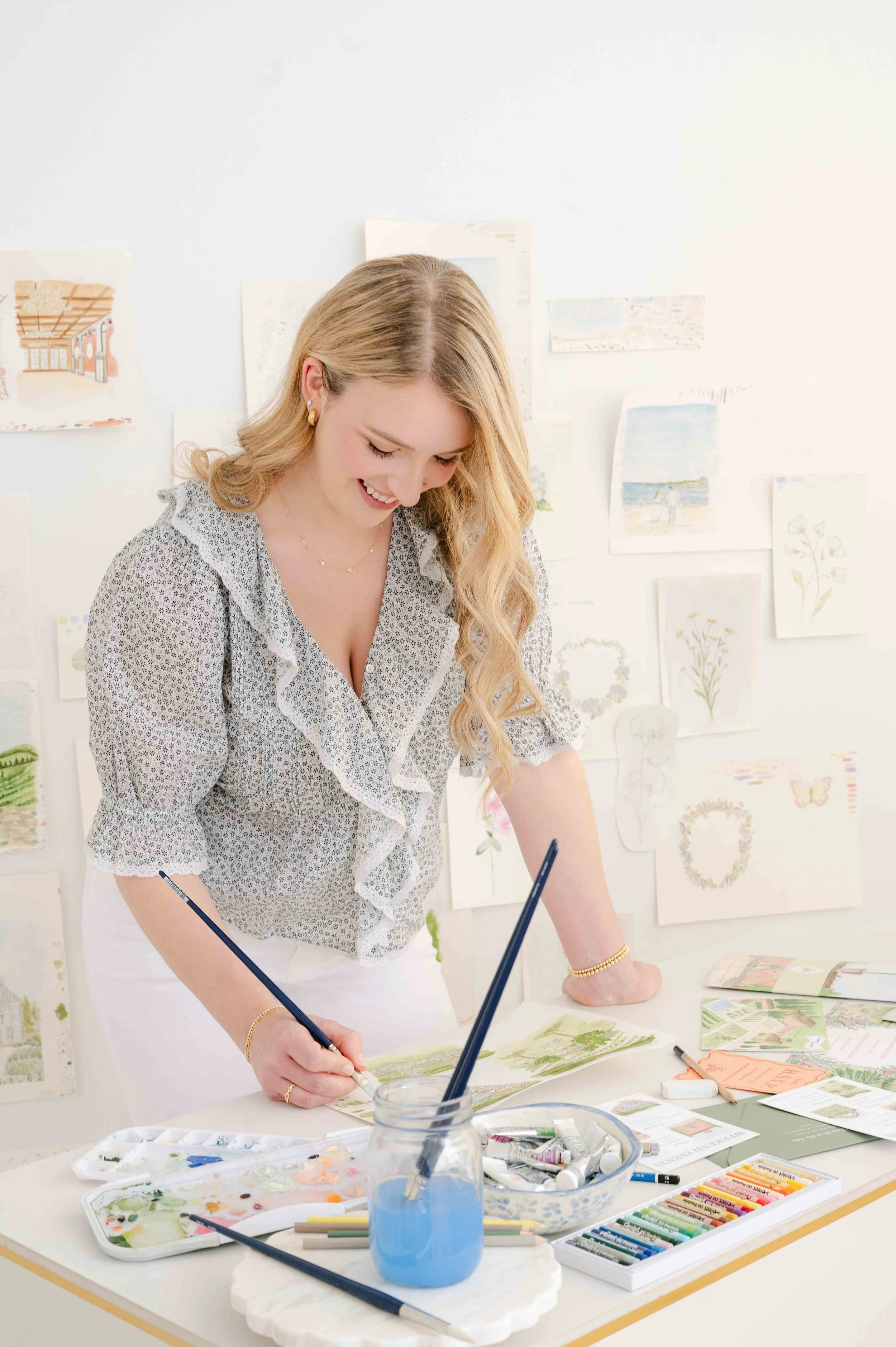 A woman smiling and painting at a desk with art supplies, in a room with watercolor paintings on the wall.