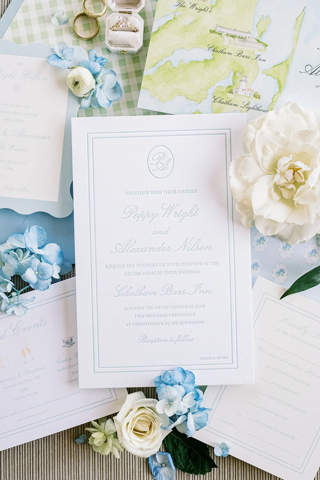 Elegant, blue wedding invitation on top of coastal-themed wedding stationery with blue and white florals around the invitation on a neutral background