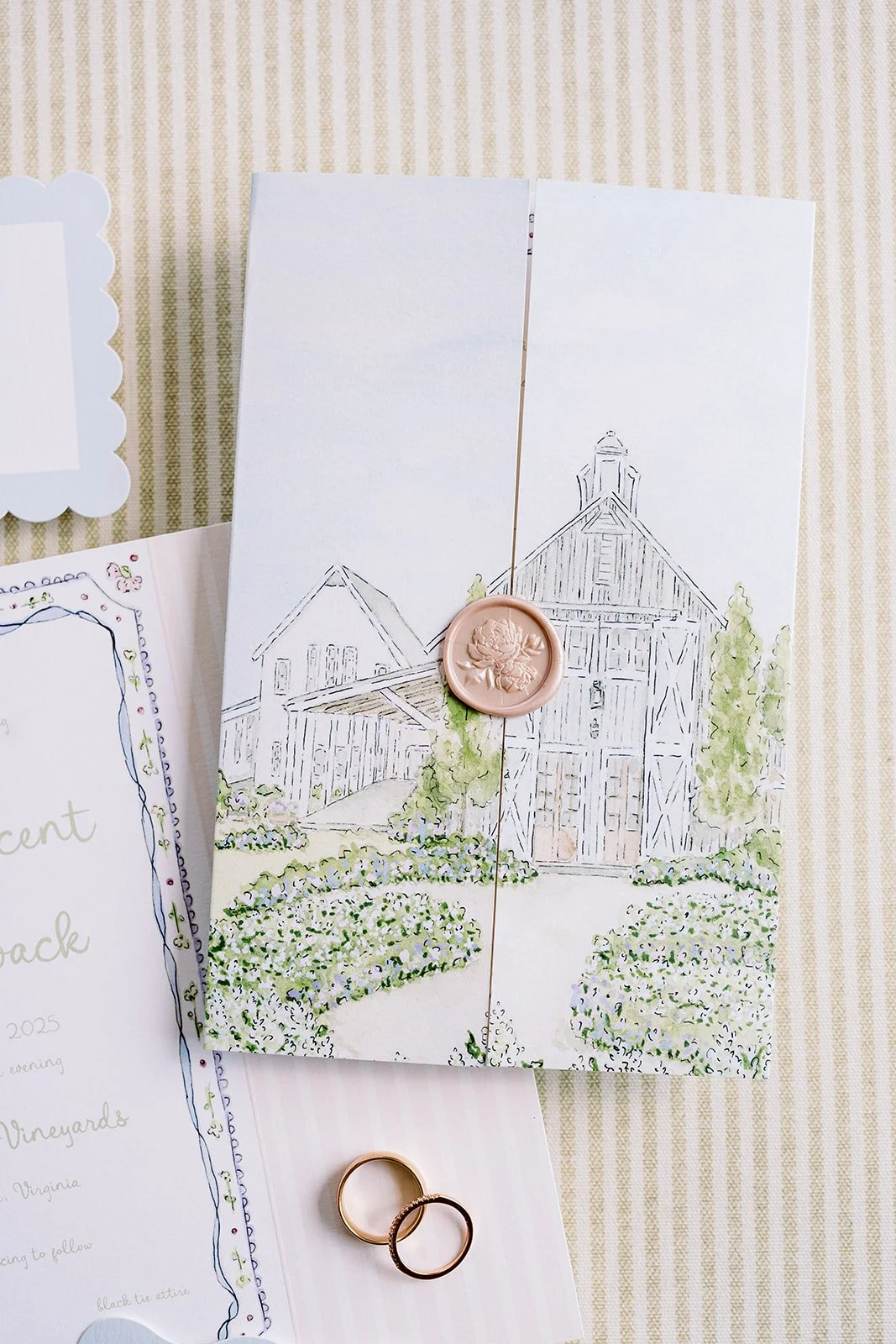 Tri-fold wedding invitation with a wedding venue watercolor illustration, sealed with a wax seal, surrounded by wedding rings, on striped background.