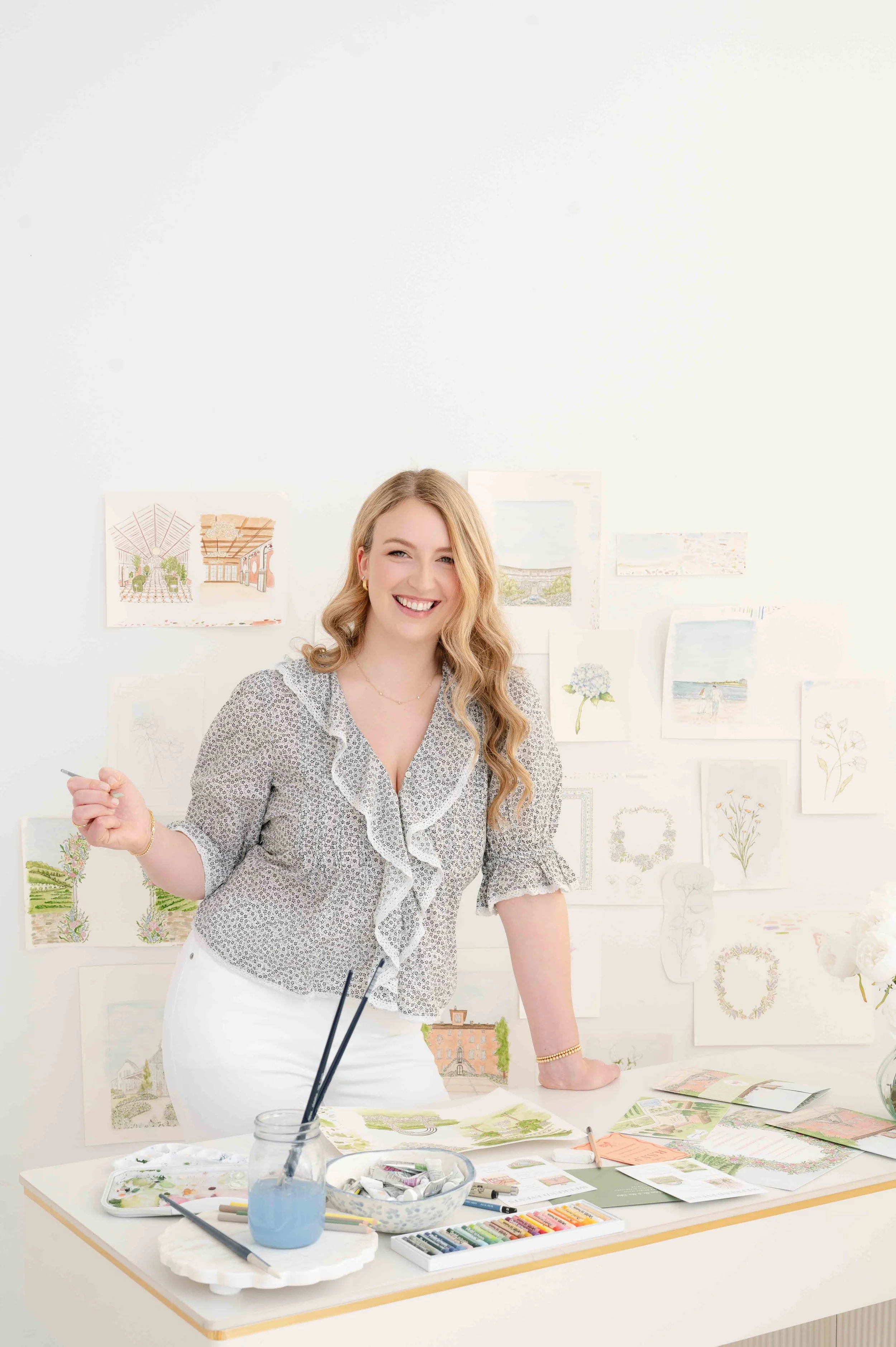 A woman with blonde hair smiling in an art studio with watercolor paintings and sketches on the walls, surrounded by art supplies on the table.