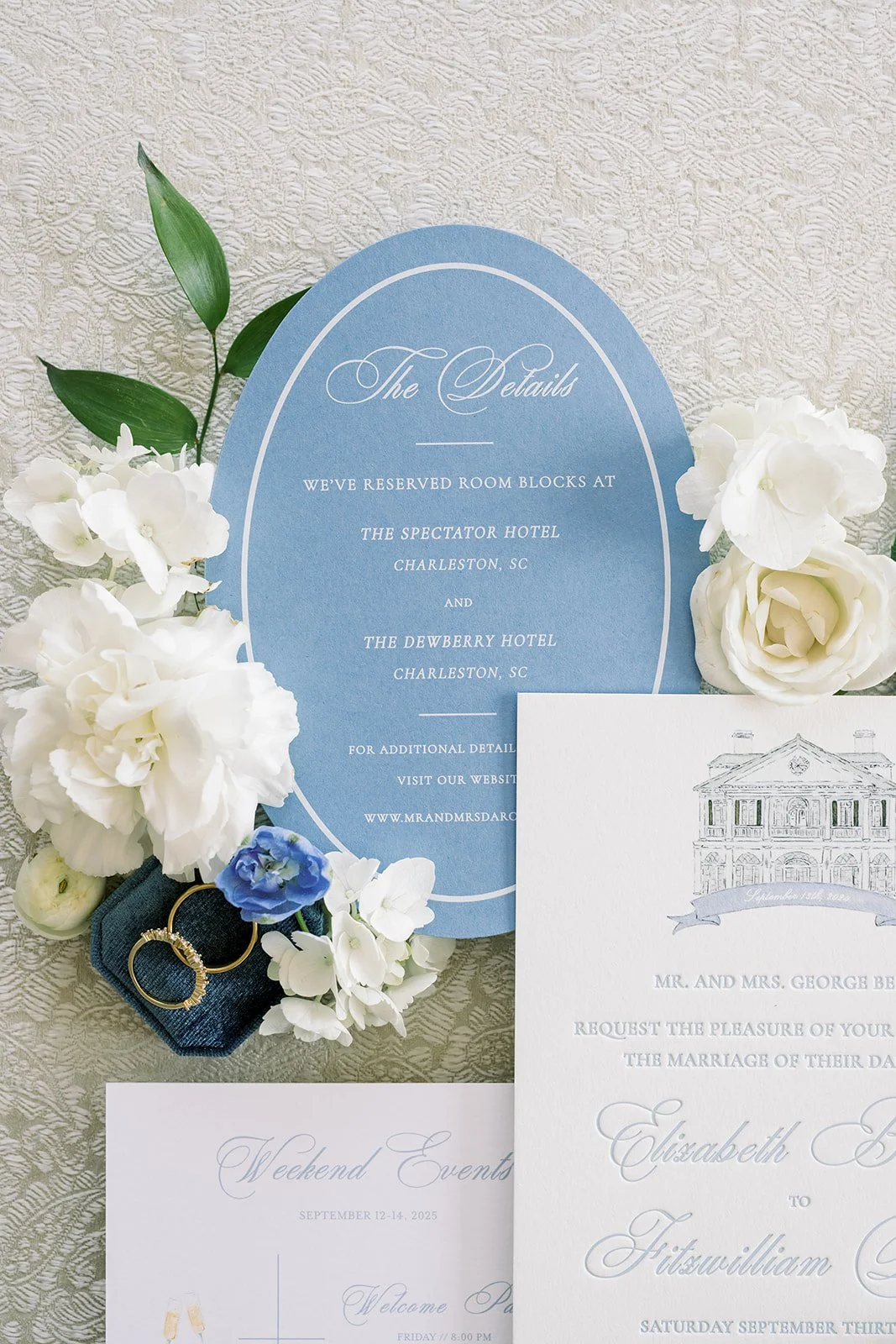 Blue oval details card next to wedding invitation and other wedding stationery with white florals and a ring
