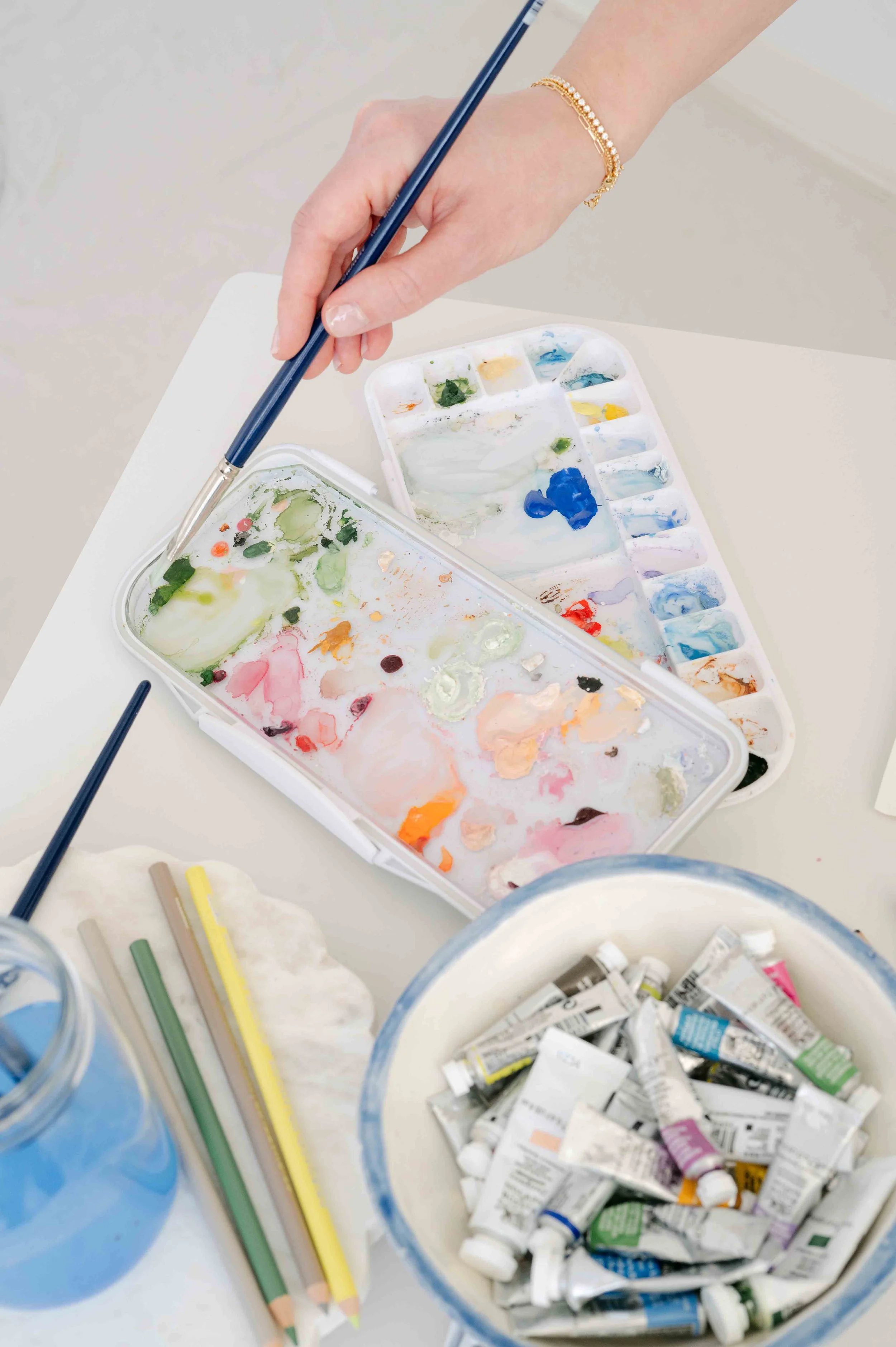Person holding a paintbrush, surrounded by watercolor palettes, tubes of paint, and colored pencils.