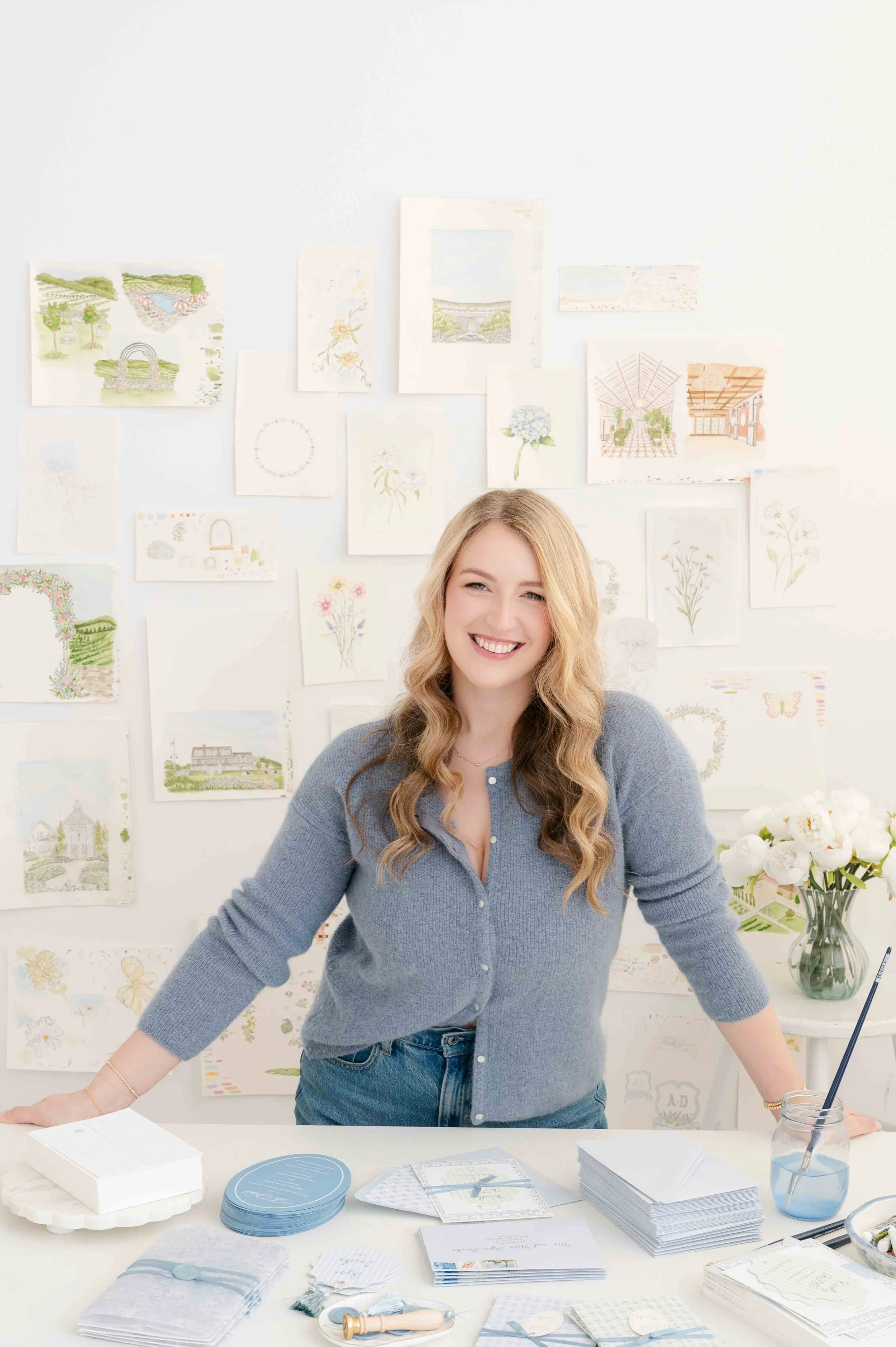 A woman with long blonde hair smiling at a desk with stationery, surrounded by watercolor paintings and drawings on the wall behind her.
