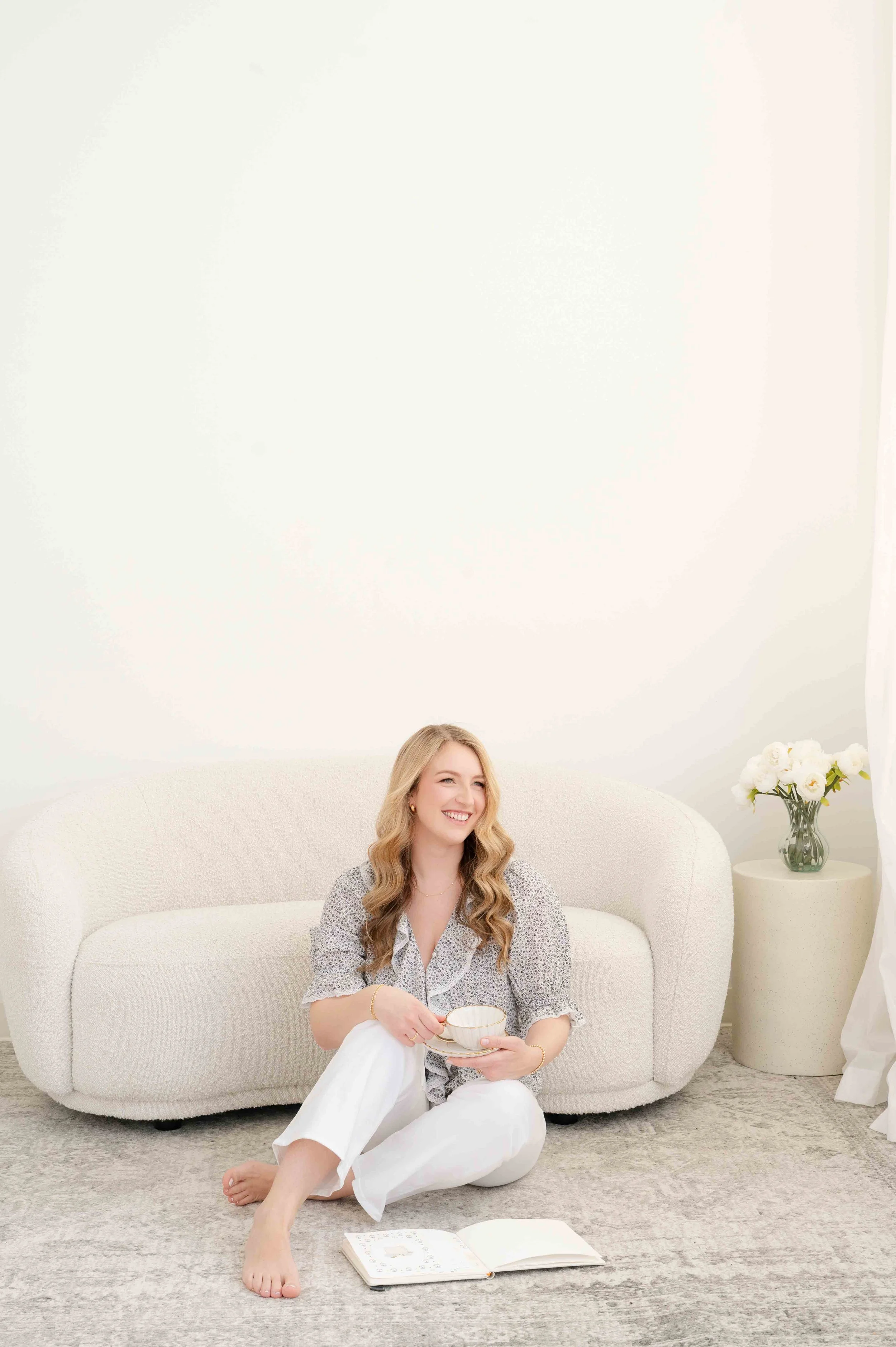 A woman with long, wavy blonde hair sitting on the floor, smiling, holding a cup, with an open book in front of her, in a bright, minimalistic living room with a cream-colored sofa and a side table with white flowers.