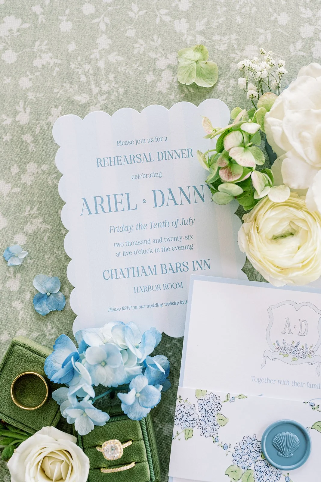 Rehearsal dinner card with fresh florals, green ring box, blue wax seal, and other wedding details on a green background