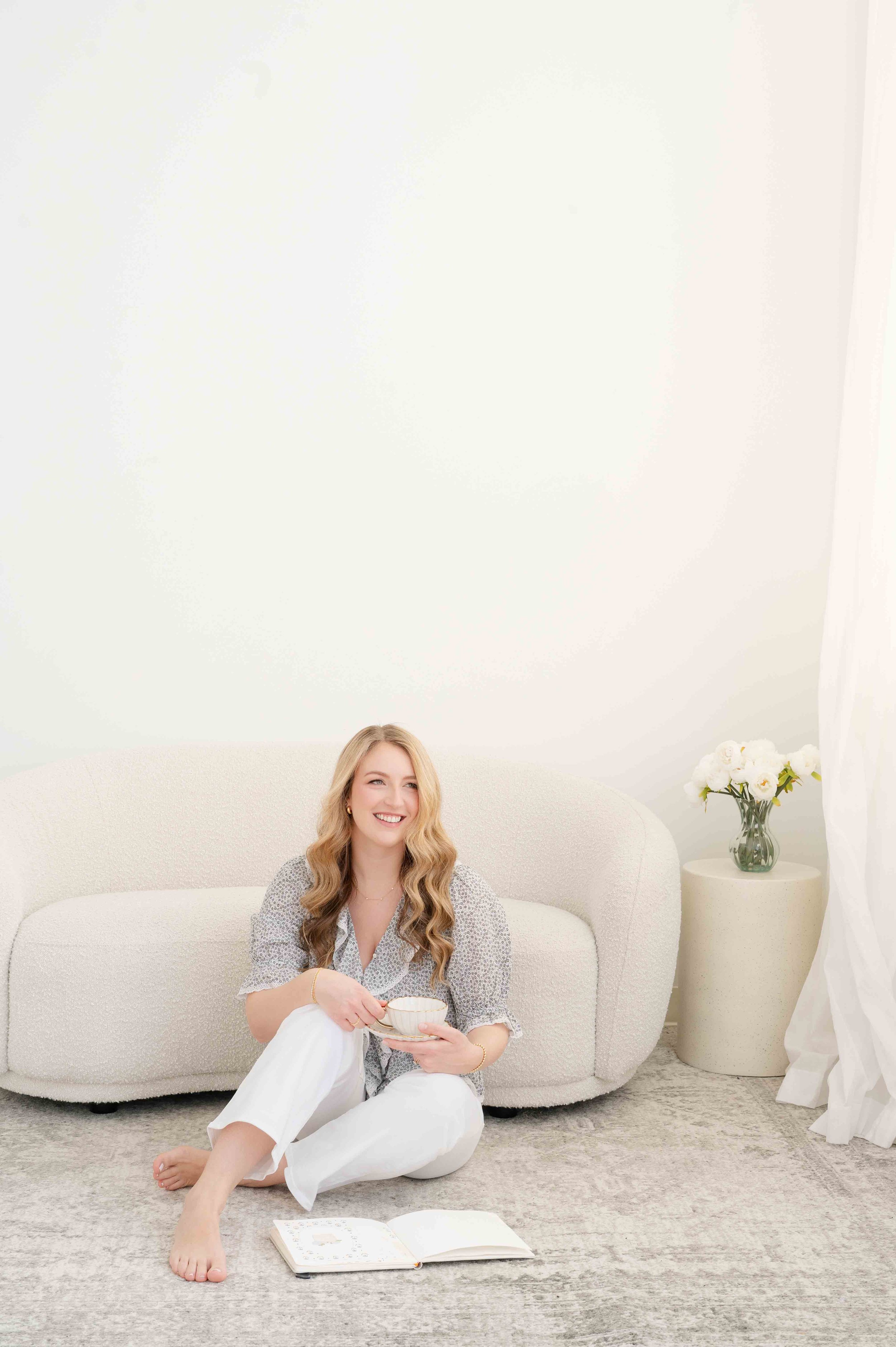 Woman sitting on the floor leaning against a couch in a neutral room with a notebook and mug