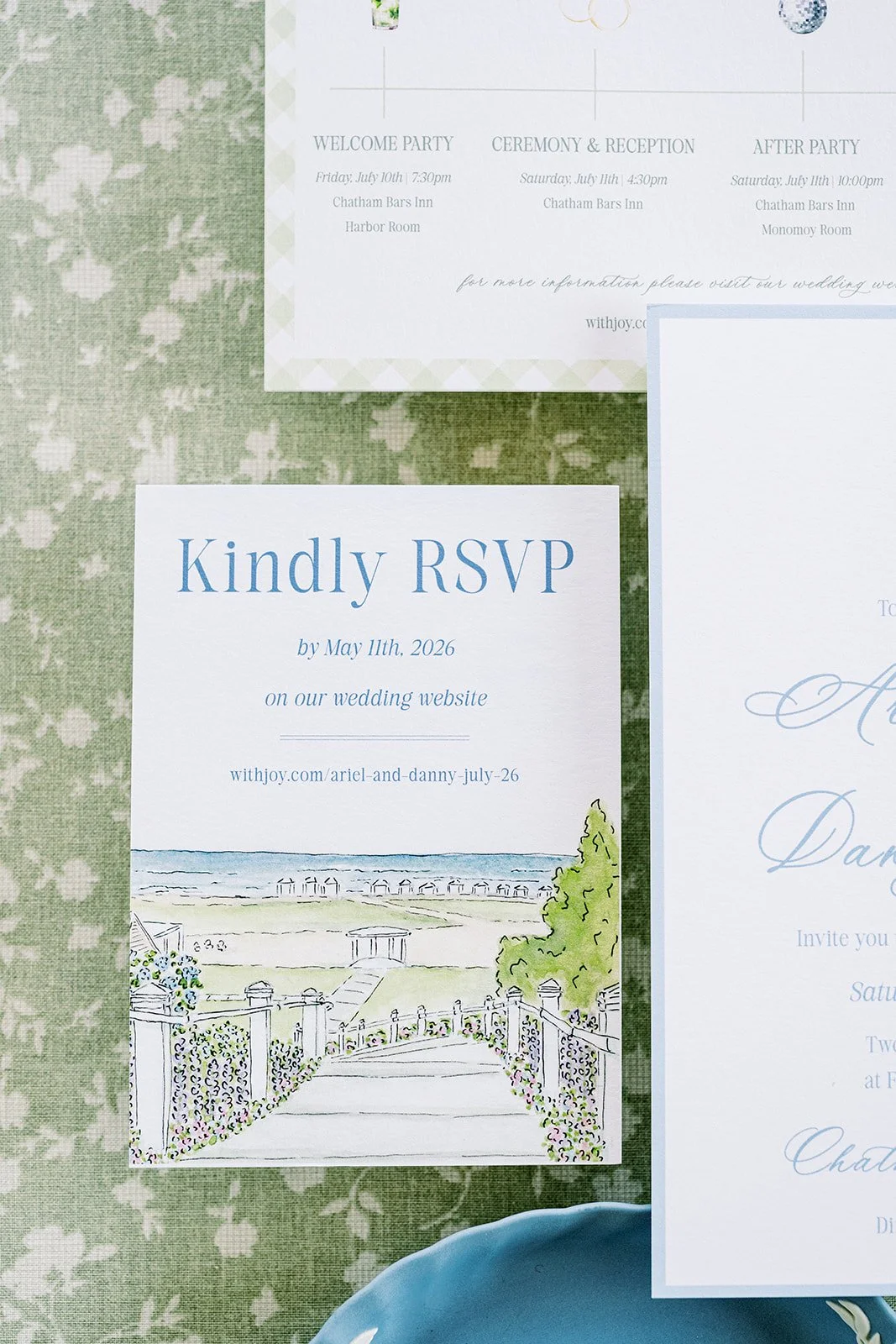 RSVP card with watercolor artwork on it next to other wedding stationery on a green floral background