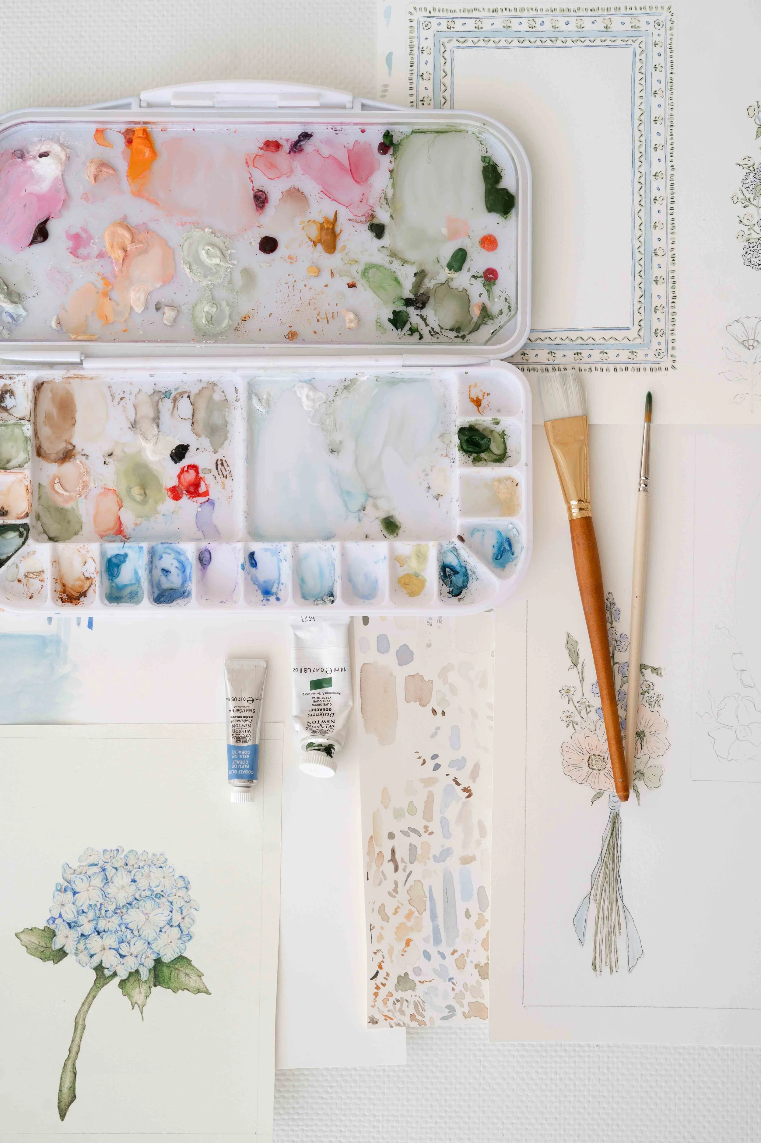 Watercolor painting supplies on a table next to floral paintings