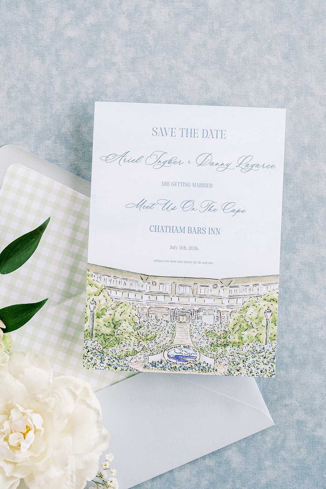 A wedding save-the-date card with a watercolor illustration featuring lush greenery and flowers next to an envelope and next to florals on a light grey background.