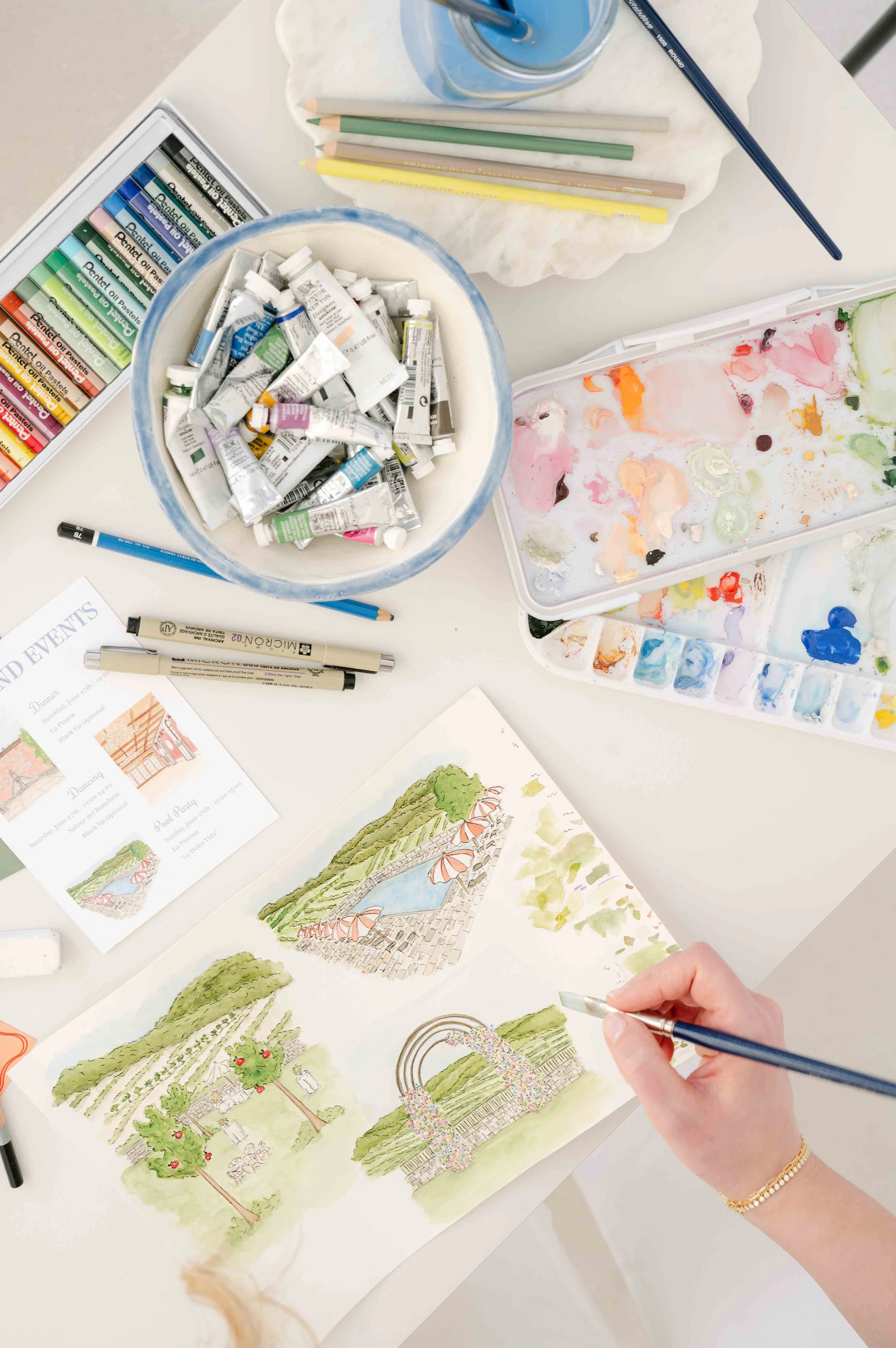 An artist's workspace with watercolor paints, sketching pens, and a hand painting a watercolor illustration of a wedding venue.