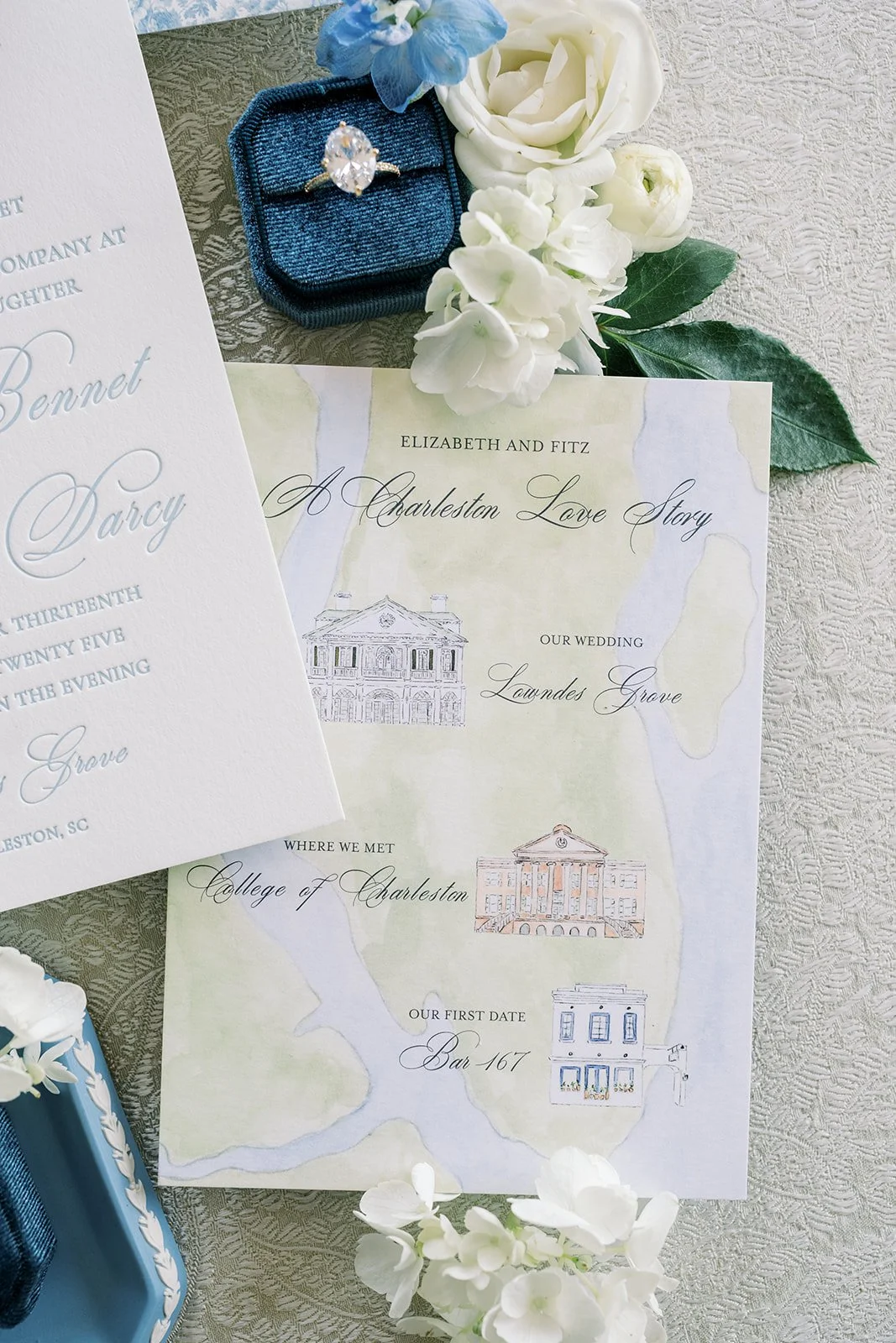 Wedding invitation with watercolor illustrations of Charleston buildings, white flowers, blue ring box with an engagement ring, and blue flower, all arranged on textured fabric.