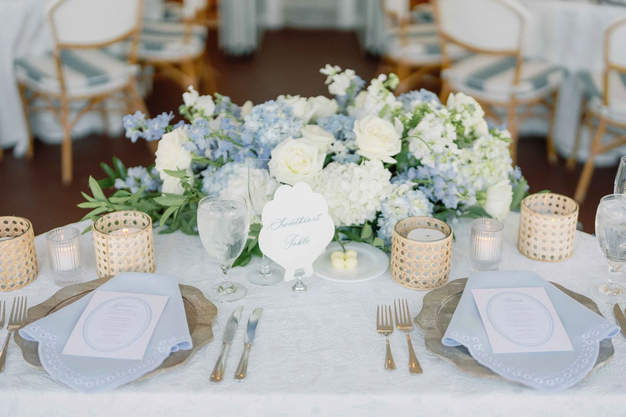 The sweetheart table featuring florals, custom menus, a shell-shaped table number, and various dinnerware in a blue and white theme