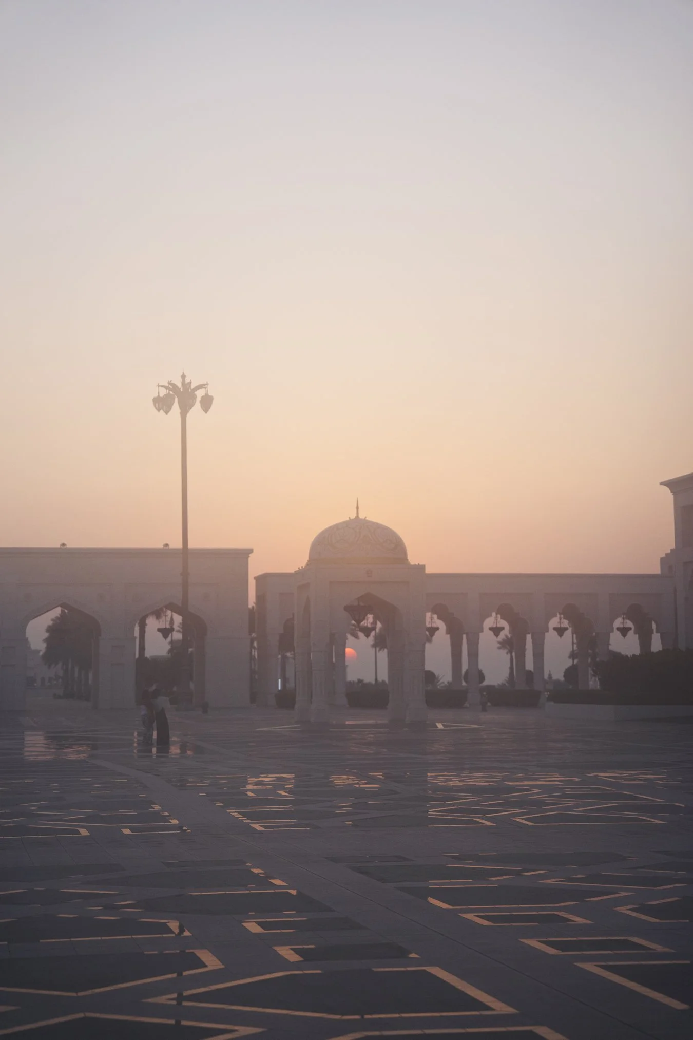 Sunset over a grand entrance with Arabic-style arches and a domed structure, illuminated by soft pink and orange hues, with a partly visible tall streetlamp and a few people walking. in Abu Dhabi UAE