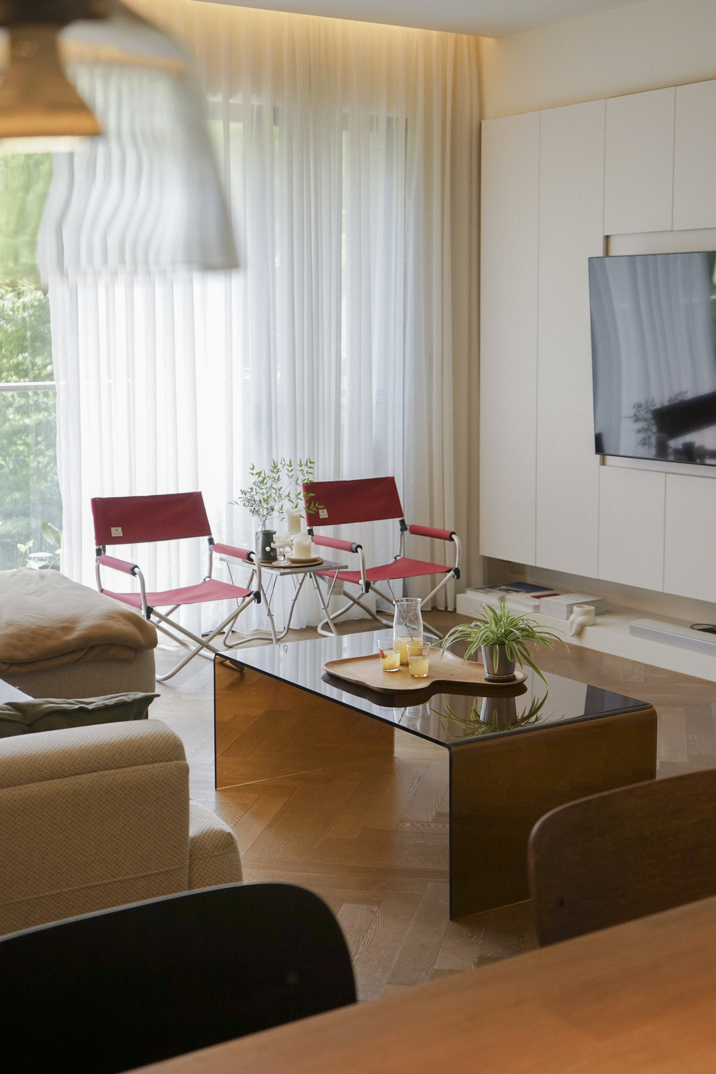 Modern living room with a black glossy coffee table holding a wooden tray with drinks and a potted plant. In the background, there are two red outdoor chairs with a small table between them. Designed by Chiang,Tsun-Yu in Teipei. Interior photography.