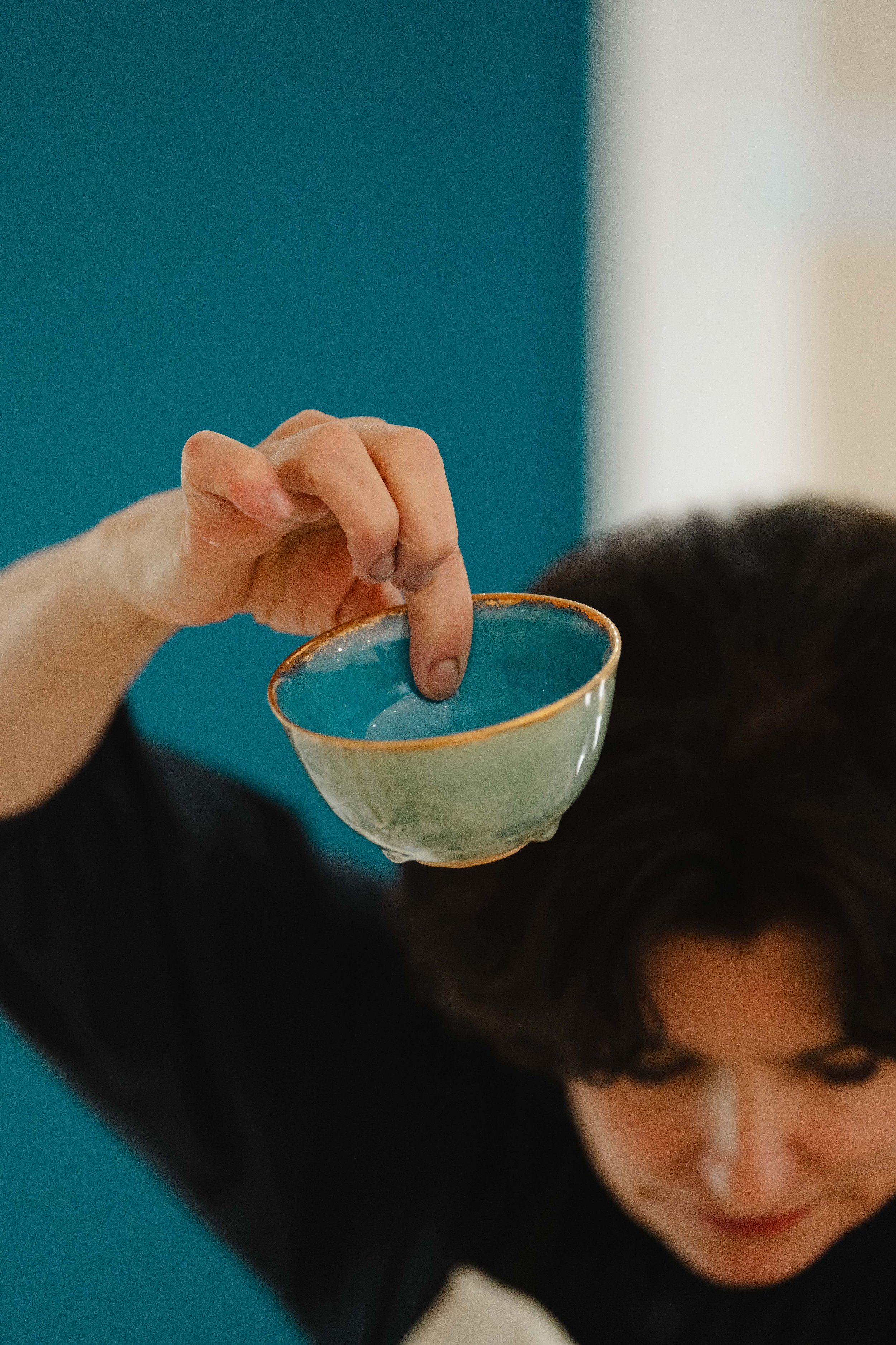 Close-up of a person holding a small ceramic bowl with a colorful glaze, pouring or dipping into water, with dark hair and a black shirt, against a blue background. Karen Swami's handmade French art ceramics. Atelier Cologne opening event in Taipei. 