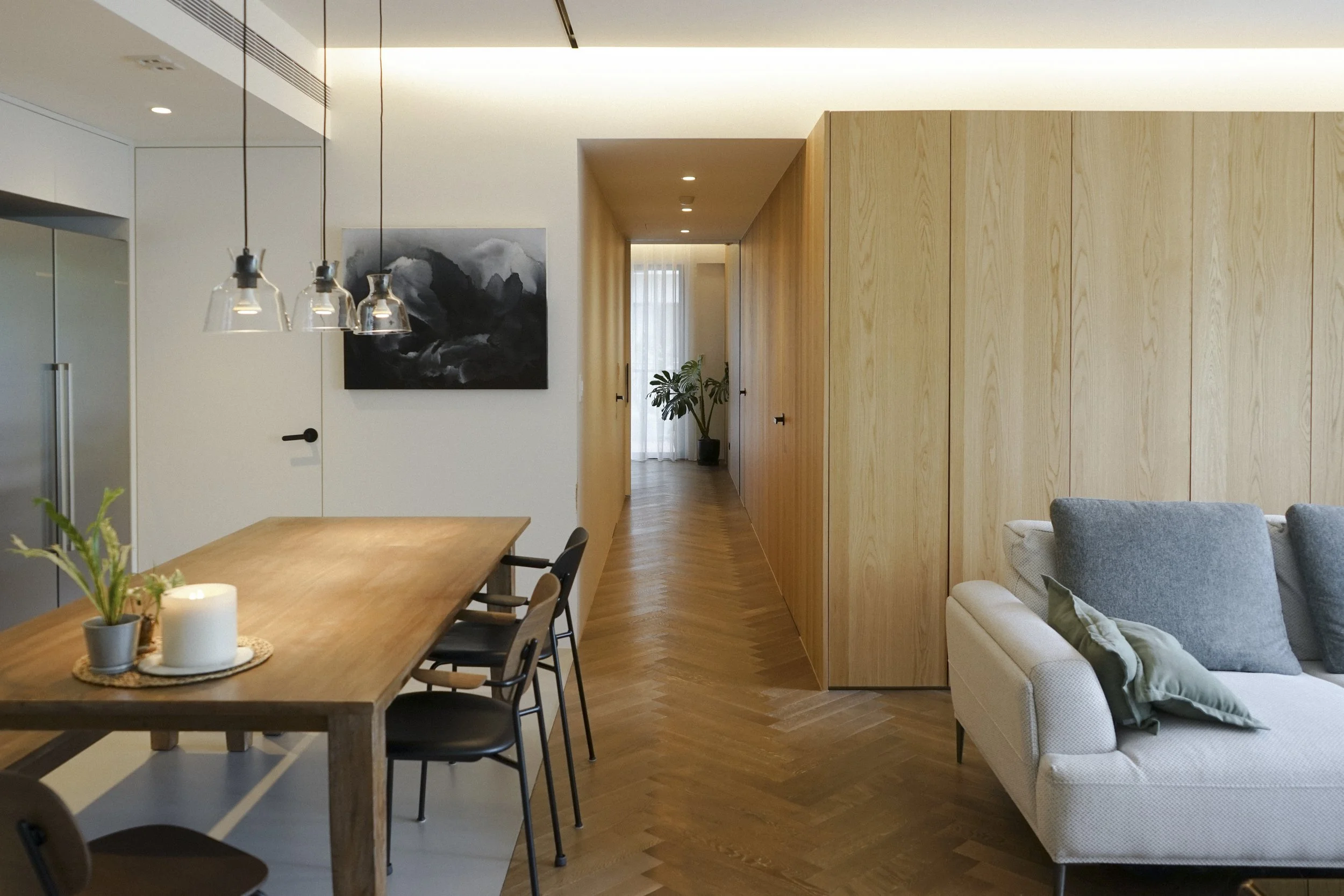Modern living room with a wooden dining table, black and gray chairs, a black and white abstract painting on the wall, wooden storage closets, and a hallway with a potted plant at the end. designed by Chiang,Tsun-Yu in Taipei. Interior photography.