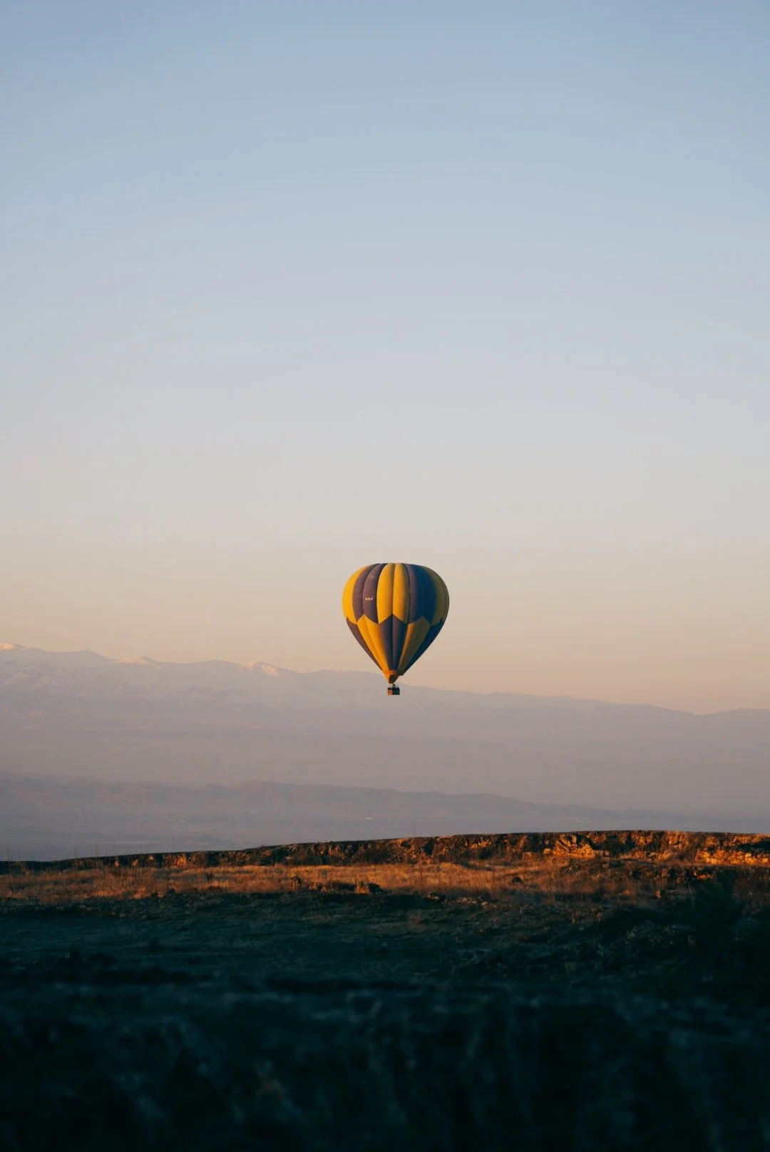 A hot air balloon with a yellow, blue, and black pattern floats in the sky above a landscape with mountains in the distance during sunrise or sunset. Pamukkale, Turkey