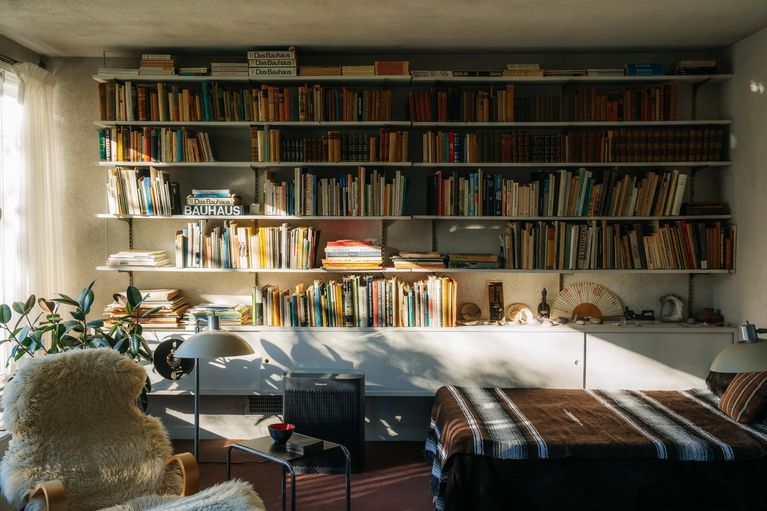 A cozy living room with a large bookshelf filled with books, including Bauhaus book. A fluffy armchair designed by Marcel Breuer, and a bed with a striped blanket. Gropius house, in Lincoln. MA. Museum, cultural space/interior photography. 