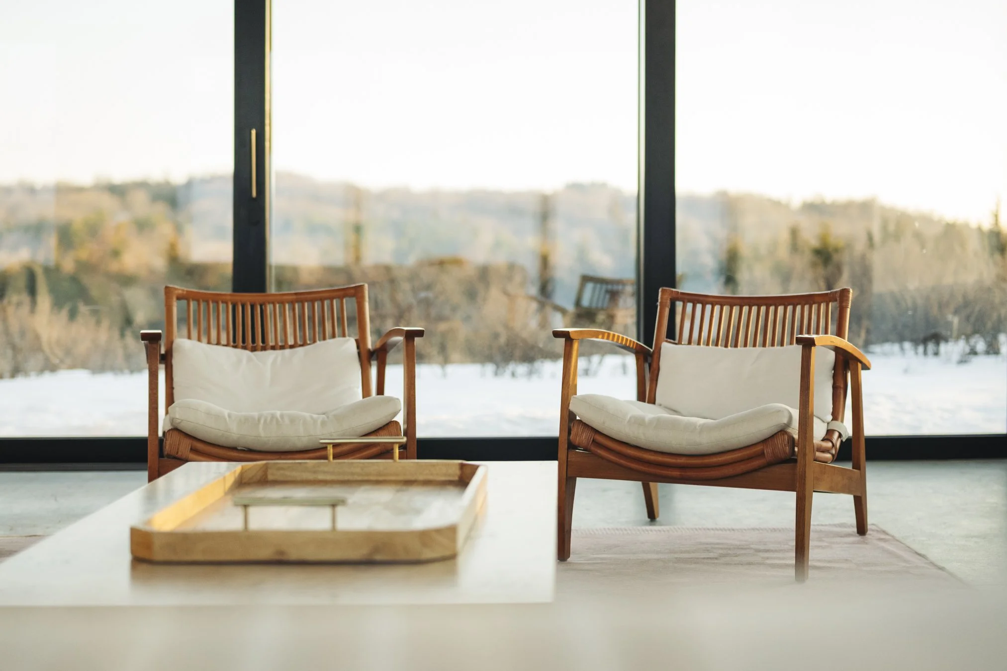 Two wooden chairs with white cushions facing a large window with a scenic outdoor view of trees and snow, and a wooden tray on a white table in the foreground. at the Taghkanic house. @redcottageincs. Interior, Airbnb photography.