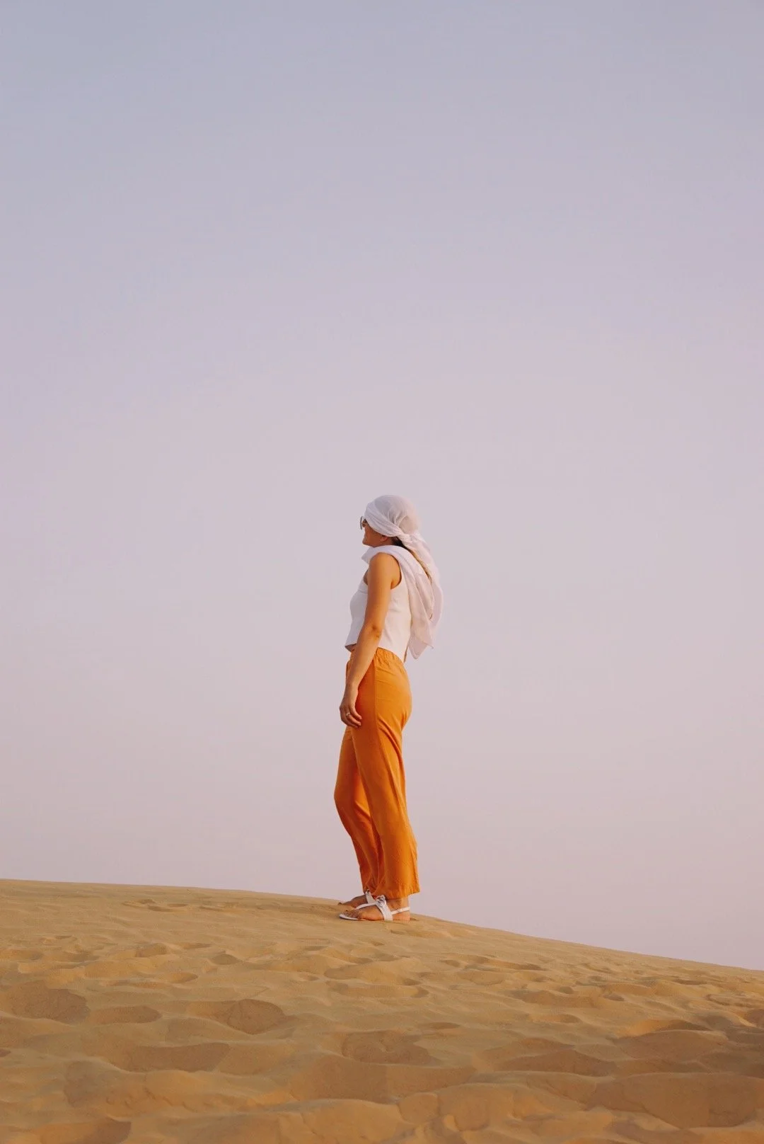 A woman standing on sand dunes, dressed in orange pants, a sleeveless white top, with a white head covering, under a clear sky. in Abu Dhabi, UAE