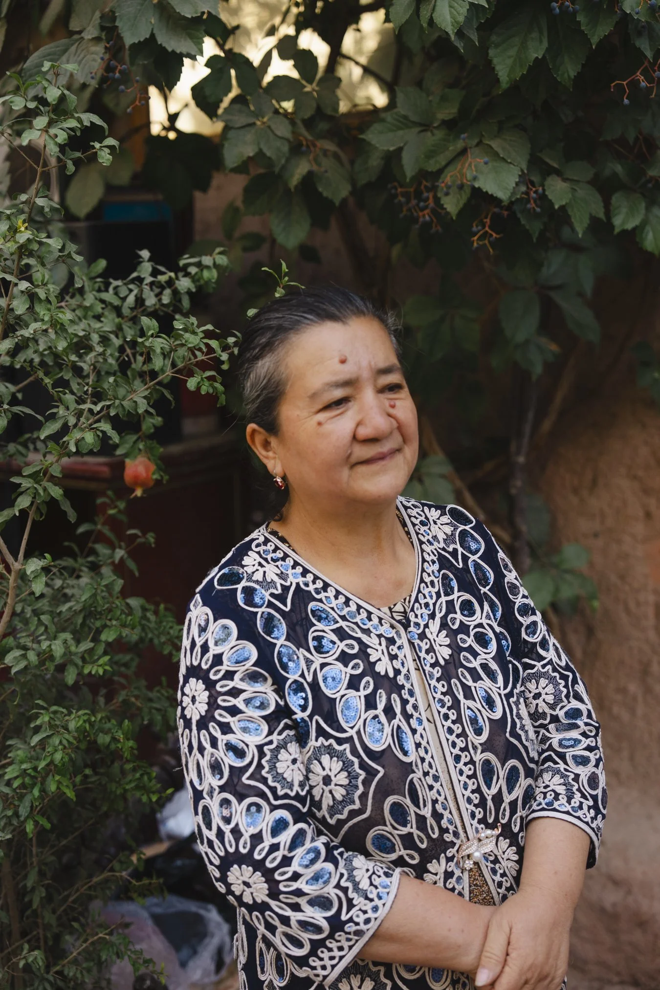 A woman with short dark hair and earrings standing outdoors among green plants and trees, wearing a black and white embroidered blouse. in Xinjiang china.
