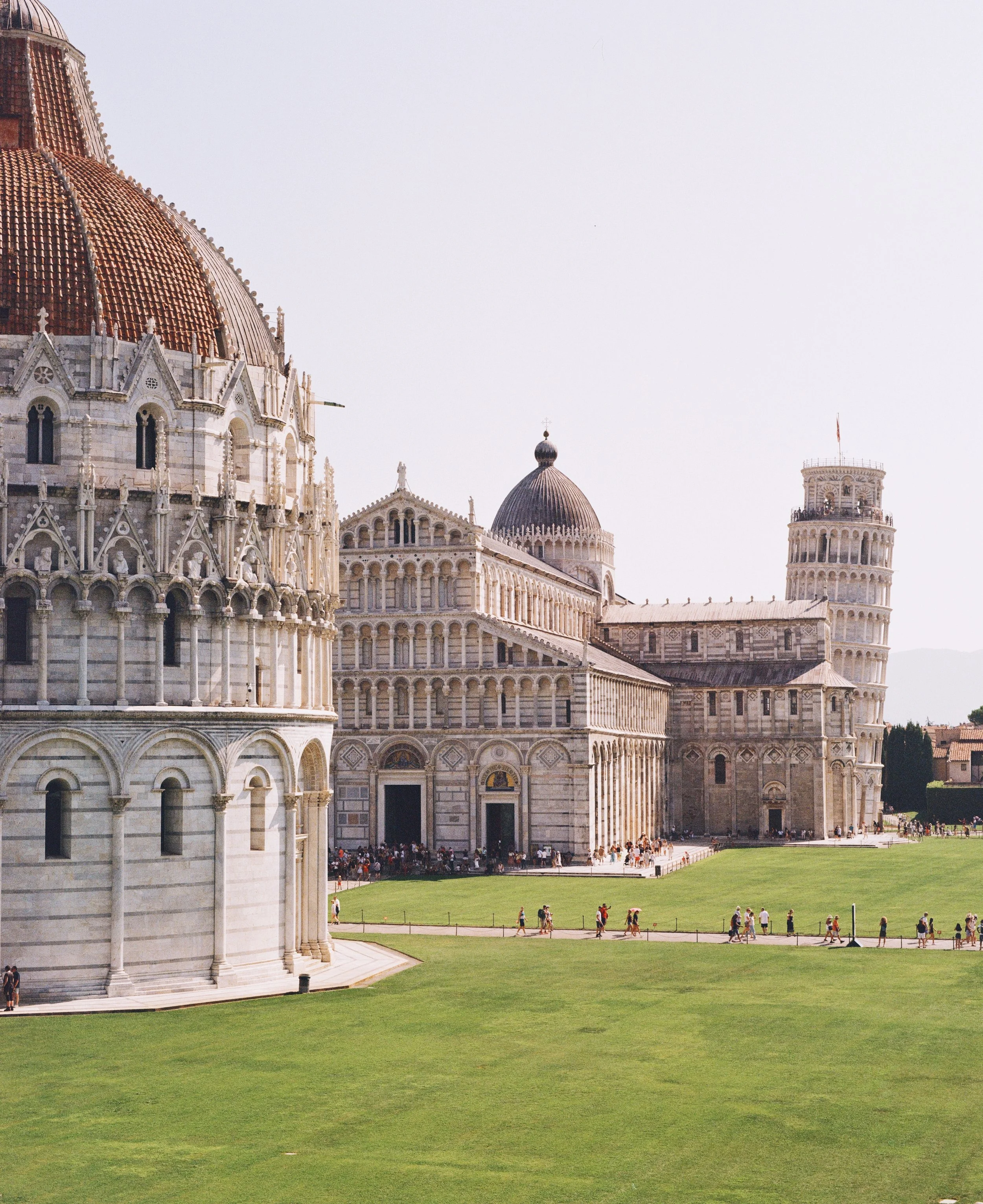 Famous Pisa Tower in Pisa, Italy, with other historic cathedral buildings, and people walking on the grass and paths.