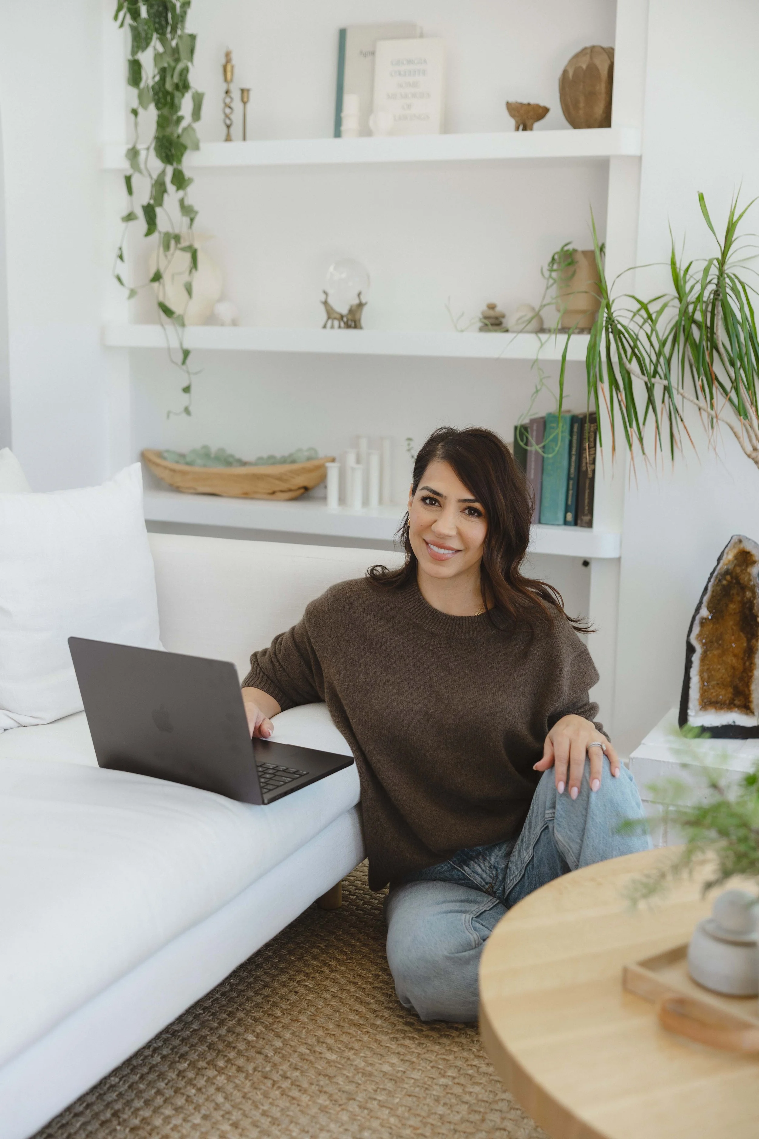 A woman with brown hair, wearing a brown sweater and jeans, sitting on the floor beside a white sofa, smiling at the camera with a laptop on the sofa arm.