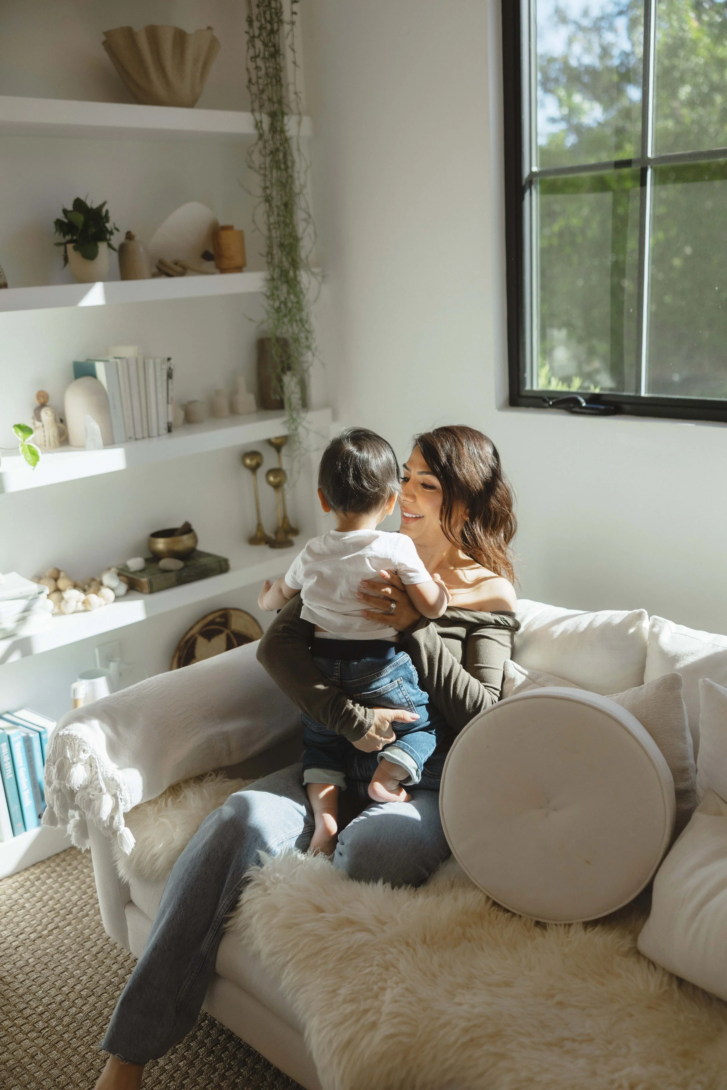 A woman sitting on a sofa holding a young child, both smiling and enjoying a moment together in a well-lit room with white walls and built-in white shelves filled with books and decorative items.