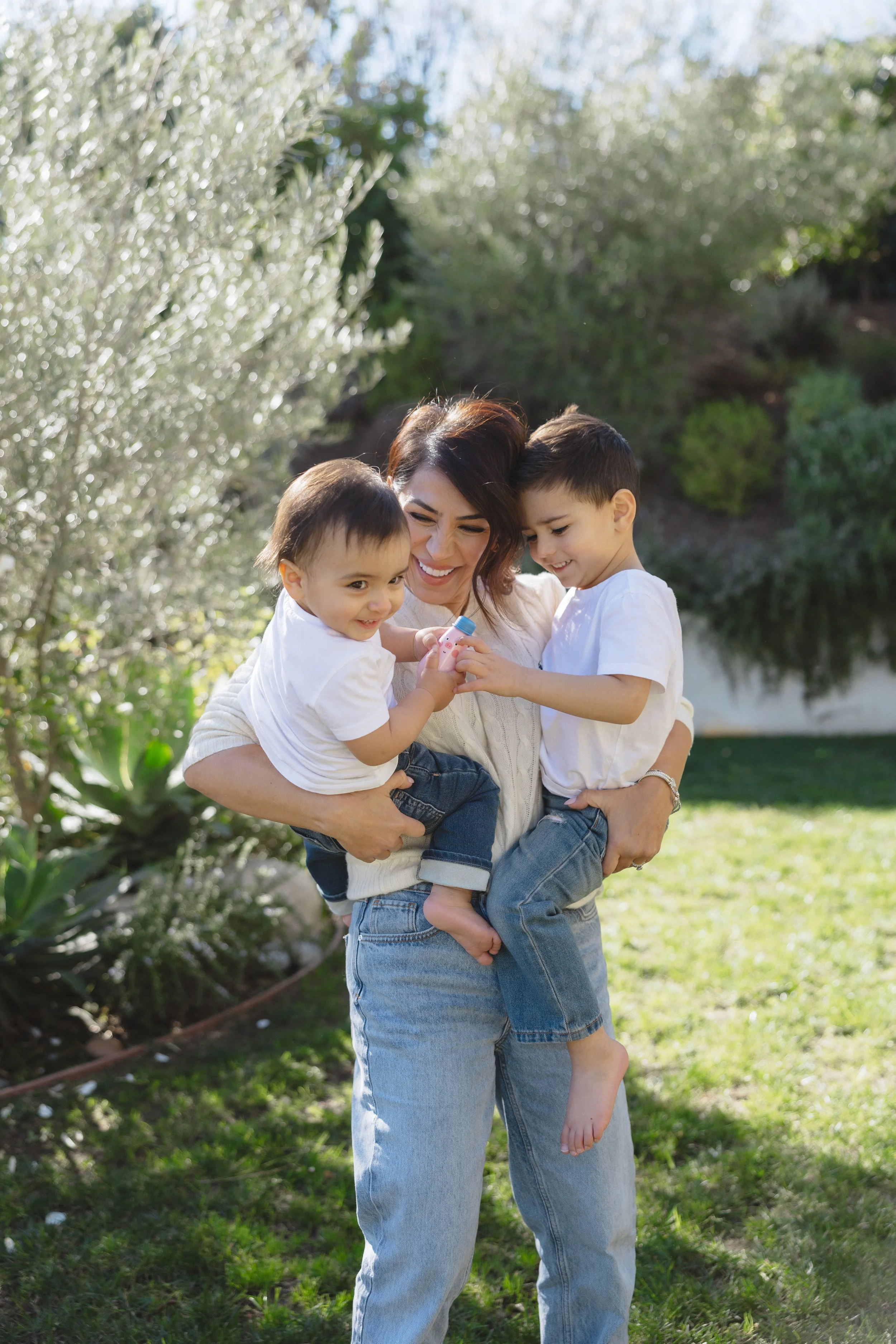A woman holding two young children outdoors in a garden, all smiling and playing with a toy.