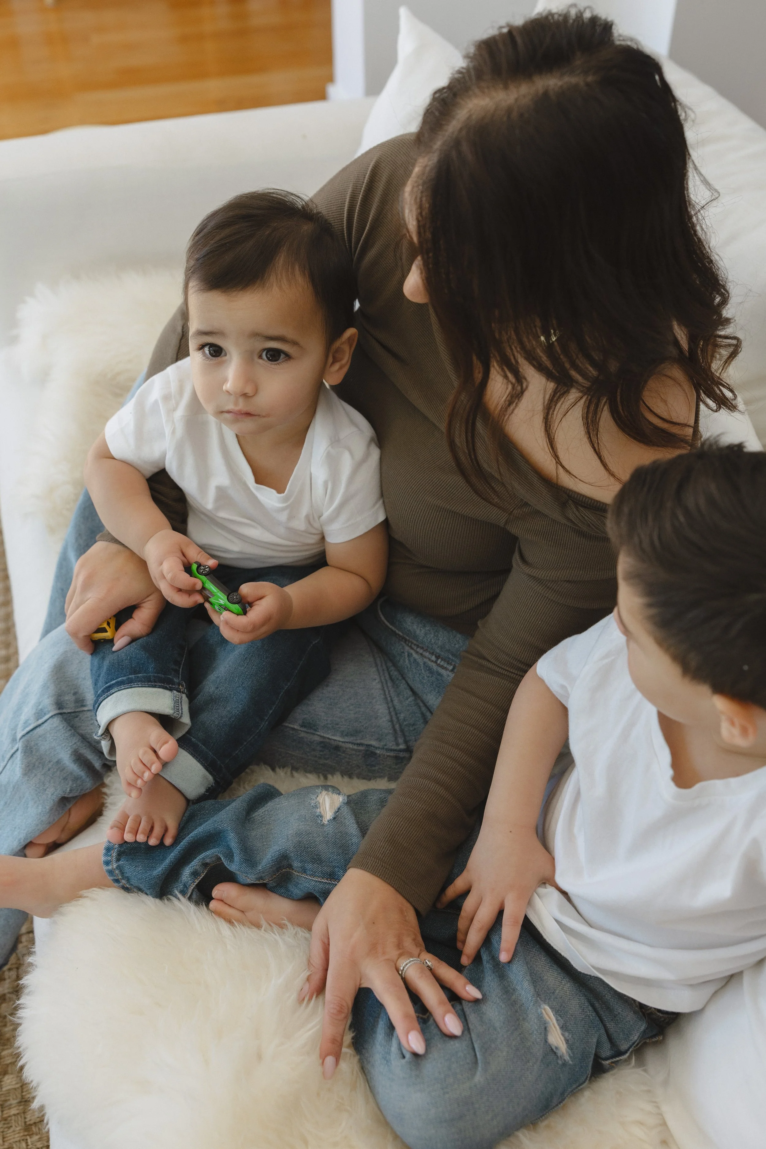 A woman with two young boys sitting on a cozy, white, furry blanket or rug on a couch or bed, having a moment of connection or conversation.