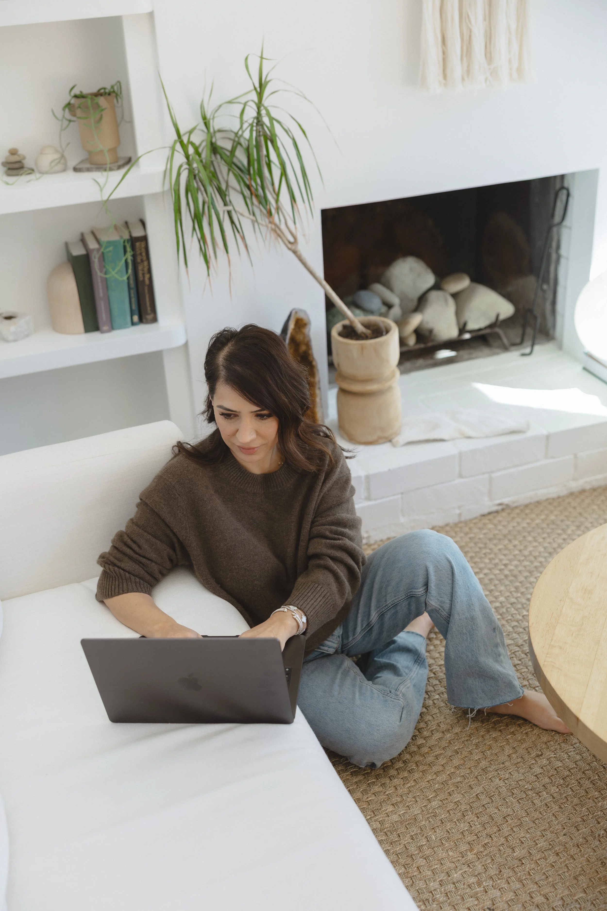A woman sitting on the floor near a white couch, working on a laptop in a cozy living room with a fireplace, potted plant, and bookshelves.