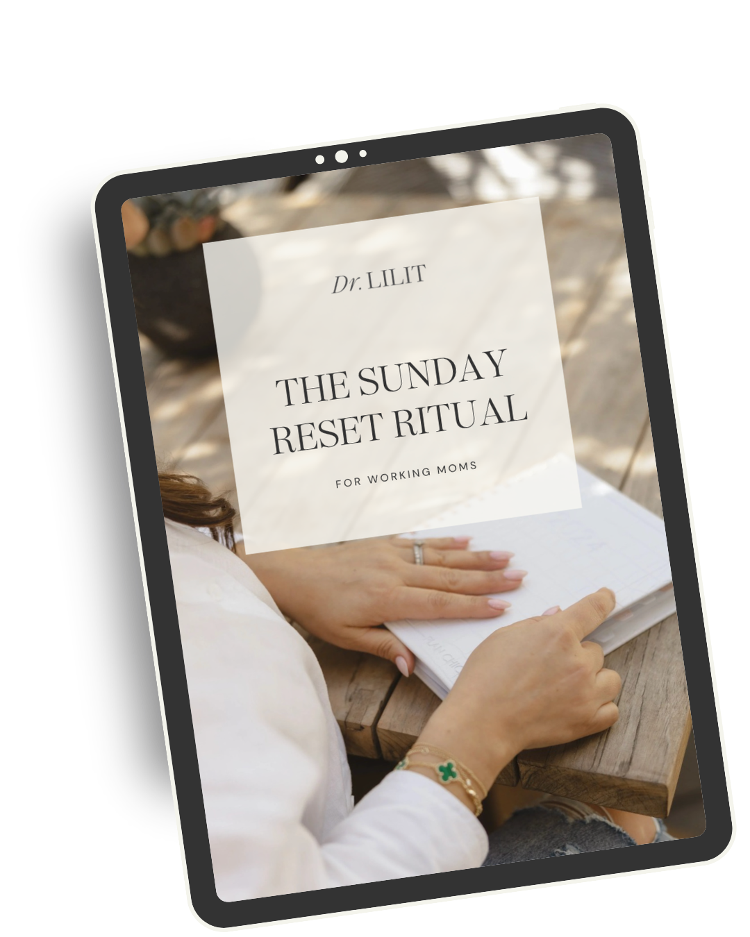 A person seated at a wooden table, holding an open notebook. The image appears on a tablet screen, with a book titled 'The Sunday Reset Ritual for Working Moms' by Dr. Lilit overlaying the scene.