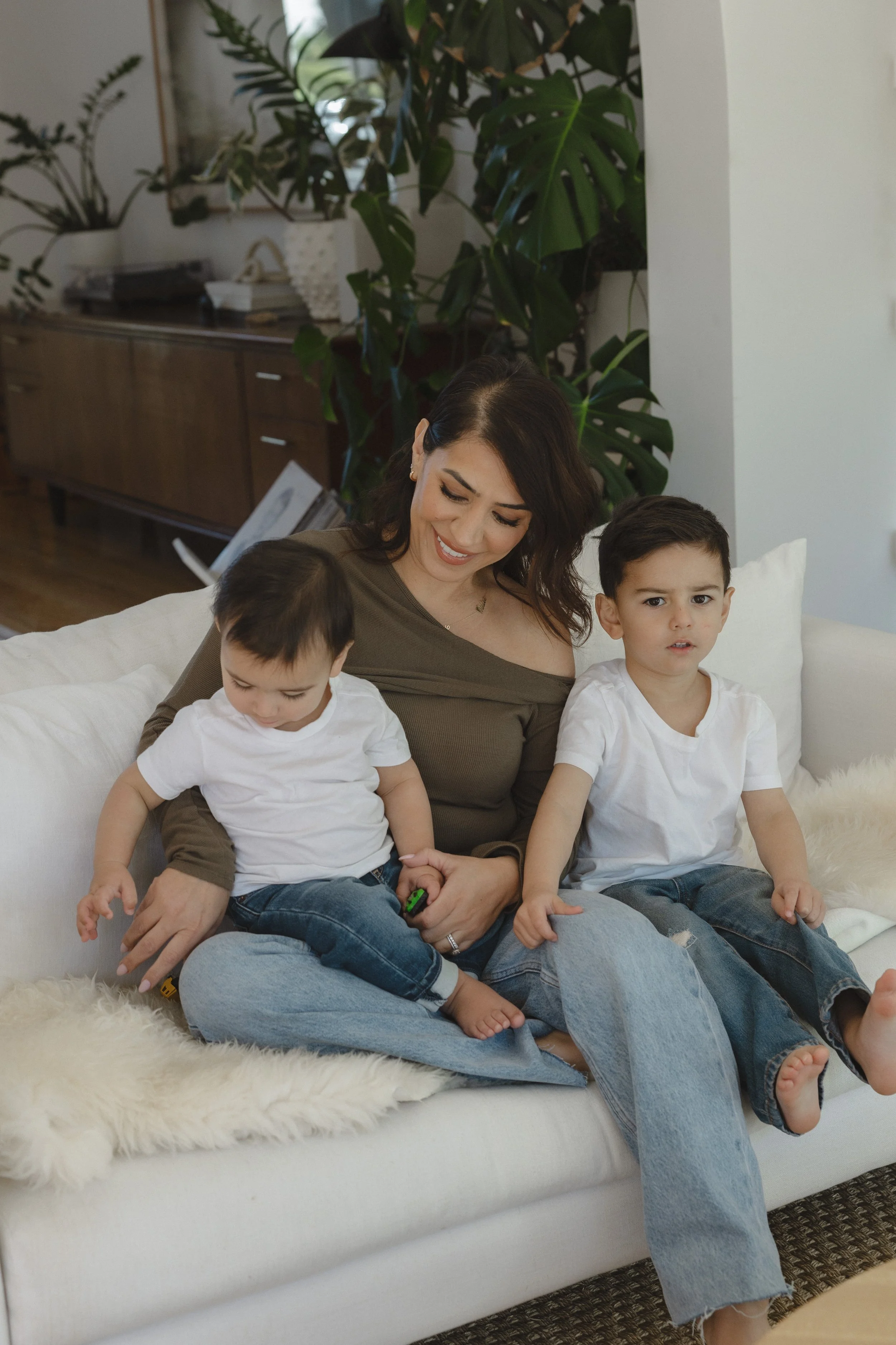 A woman sitting on a white couch with two young children, one on her lap and one next to her, in a cozy living room with plants and wooden furniture in the background.