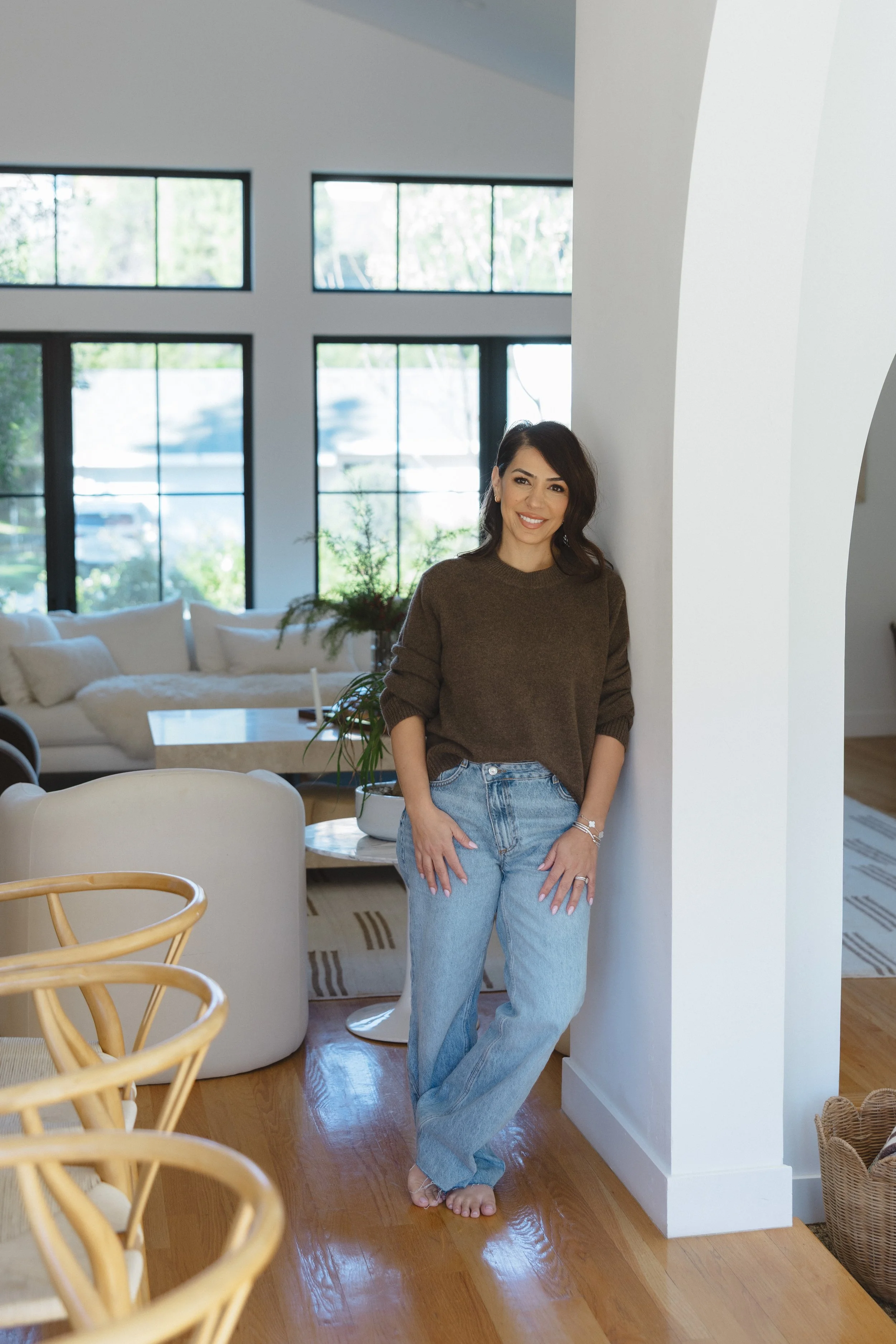 A woman standing barefoot in a modern, bright living room, smiling and leaning against a white wall.