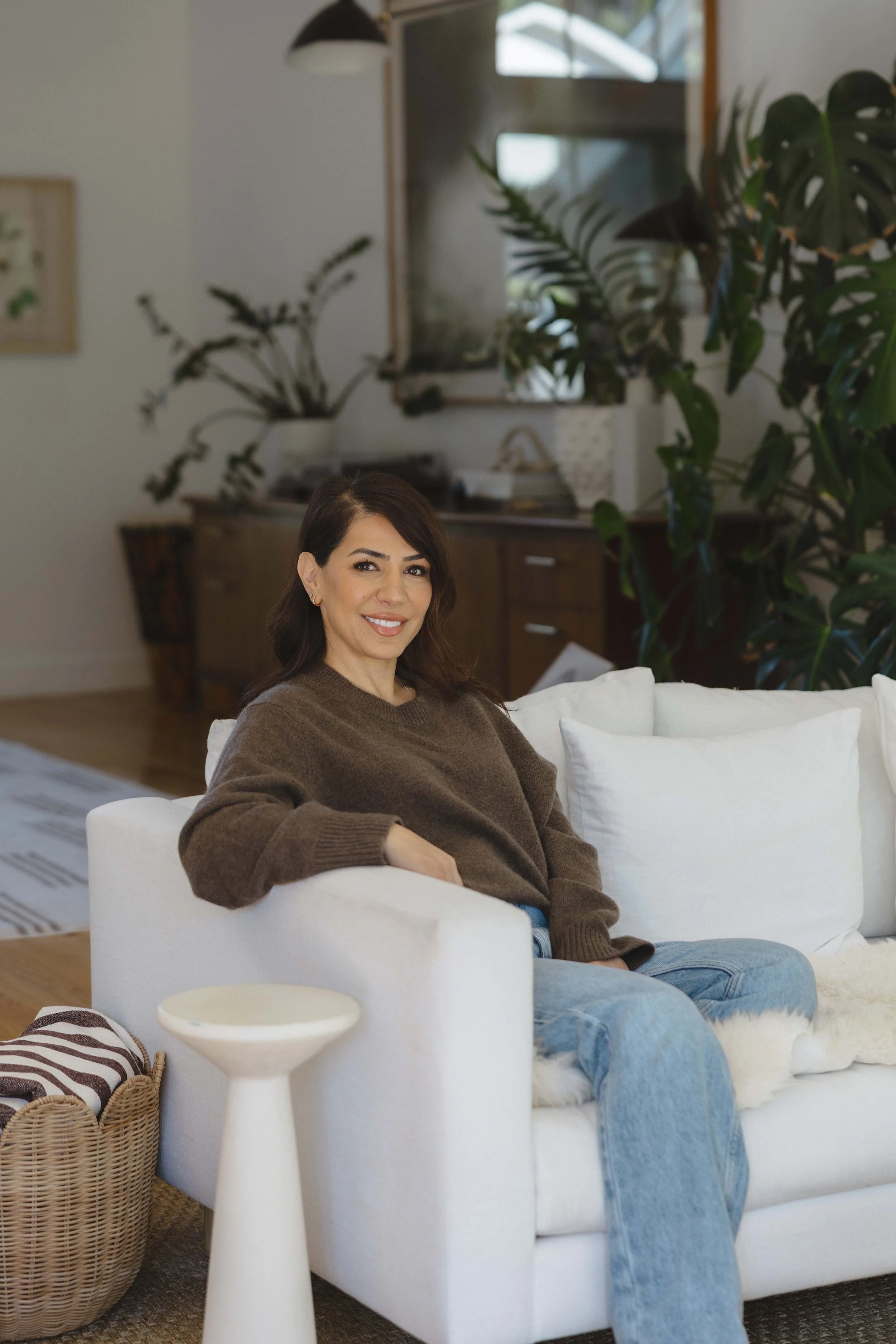 A woman with dark brown hair sitting on a white couch in a cozy living room, smiling at the camera. She is wearing a brown sweater and light blue jeans. Behind her are houseplants and a wooden cabinet.