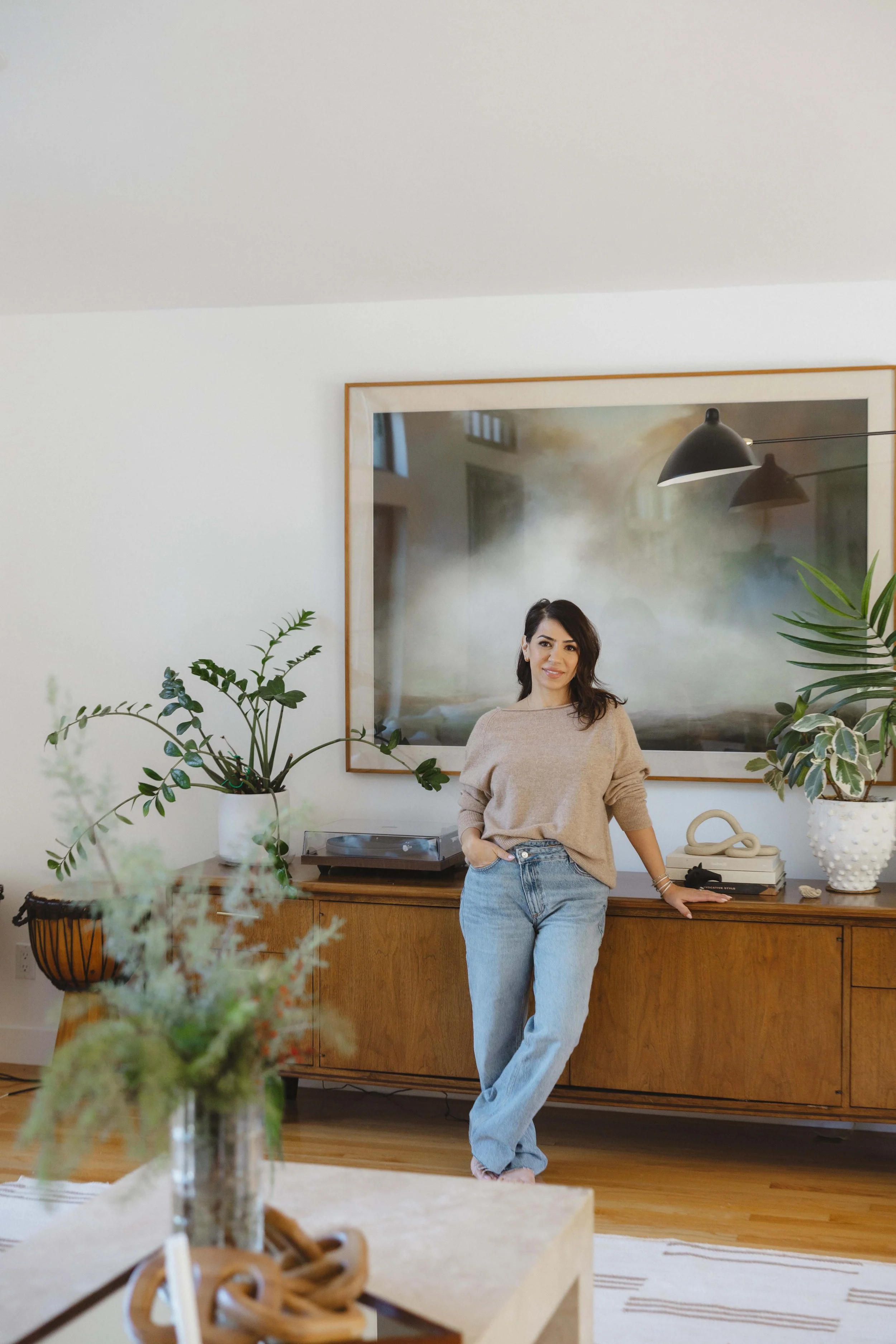 Woman standing in living room with wooden sideboard, potted plants, large framed artwork, and modern decor.