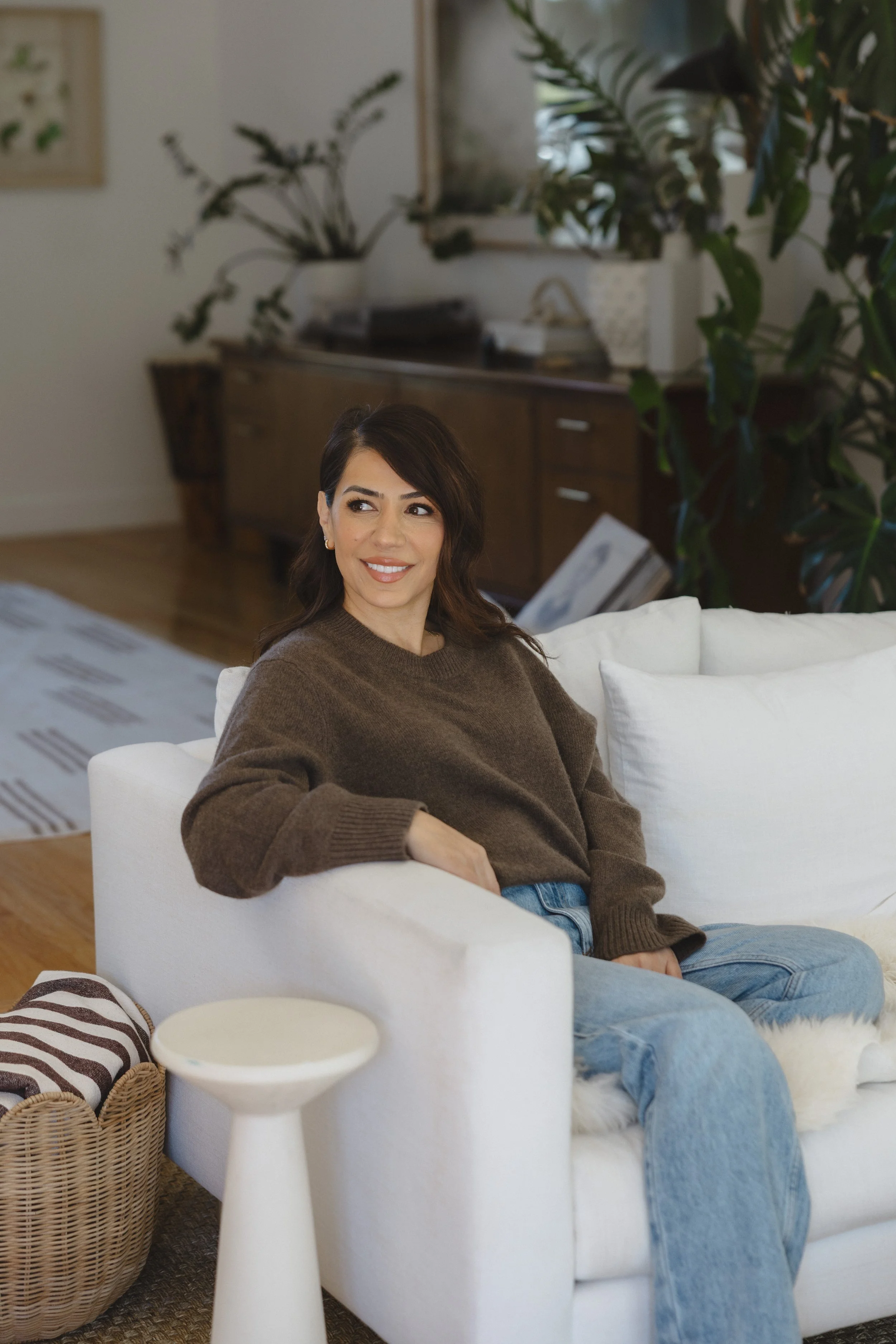 A woman with dark hair, wearing a brown sweater and jeans, sitting on a white sofa in a living room with wood flooring and green plants.
