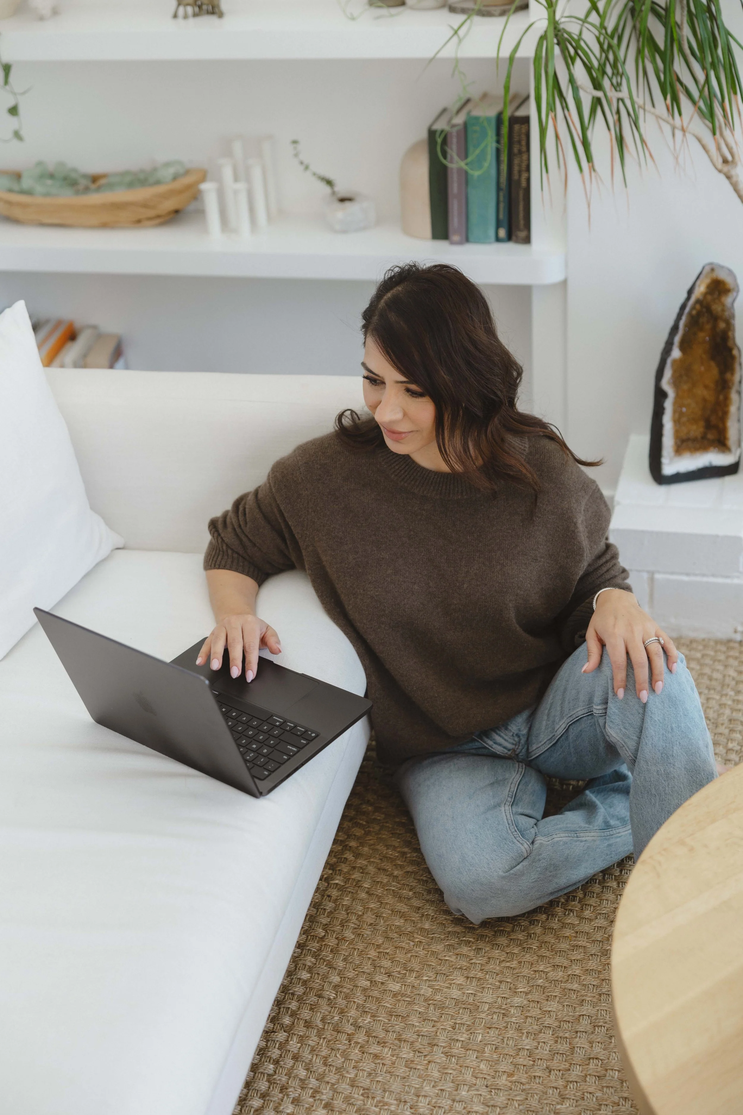 A woman sitting on the floor beside a white couch, working on a laptop, in a cozy living room with books and decorative objects.