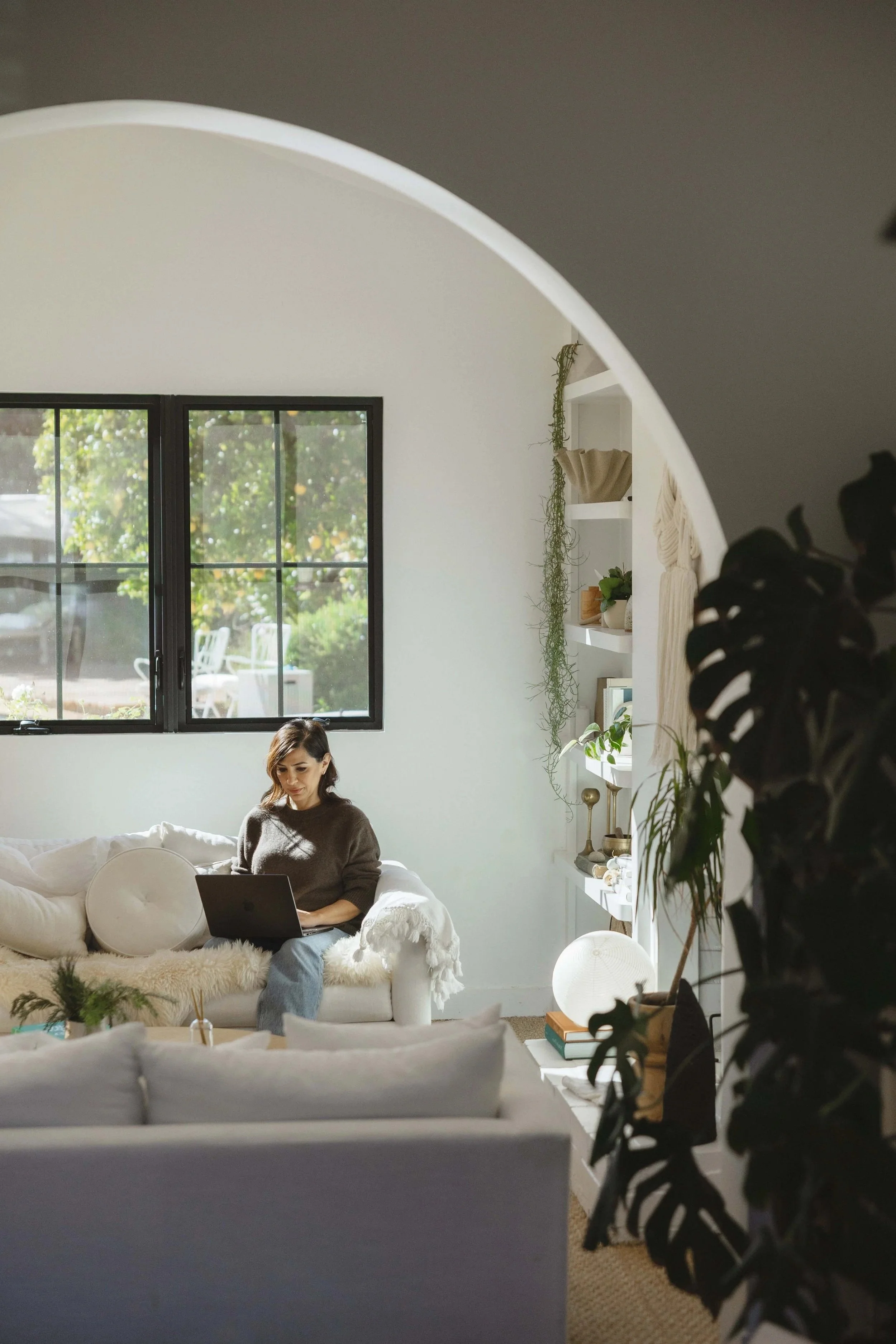 Woman sitting on a white sofa working on a laptop in a bright living room with large window and decorative plants.
