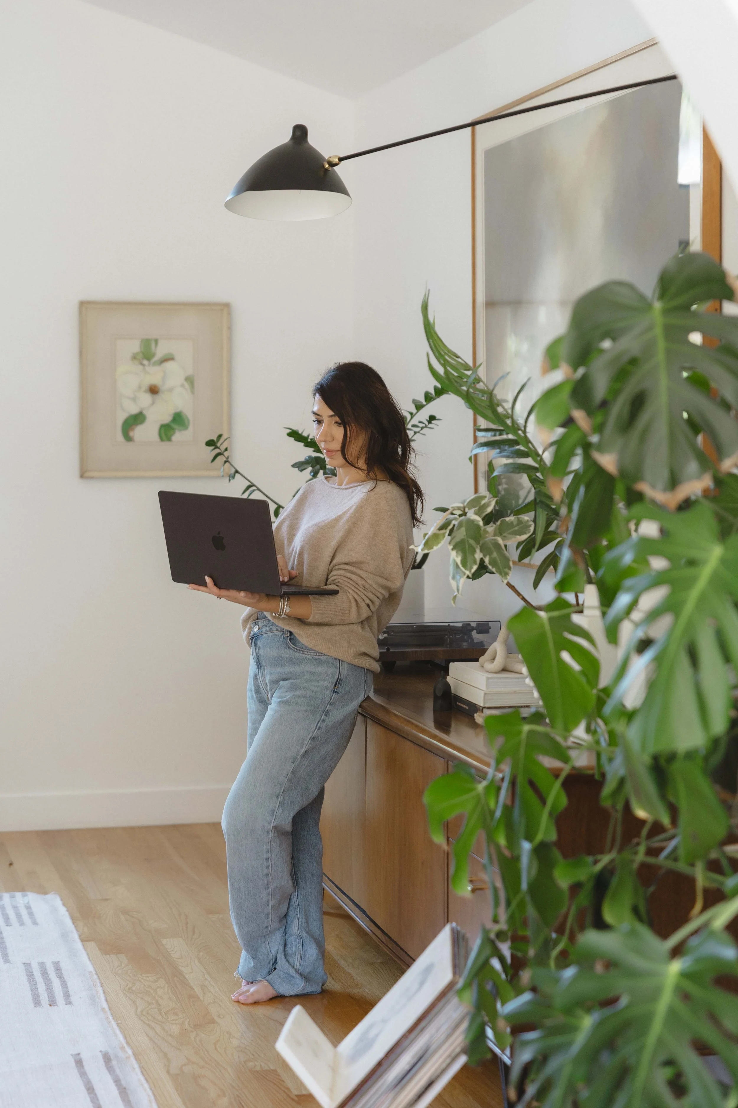 A woman with dark hair, wearing a beige sweater and blue jeans, standing barefoot in a room, using a laptop, with houseplants and artwork in the background.