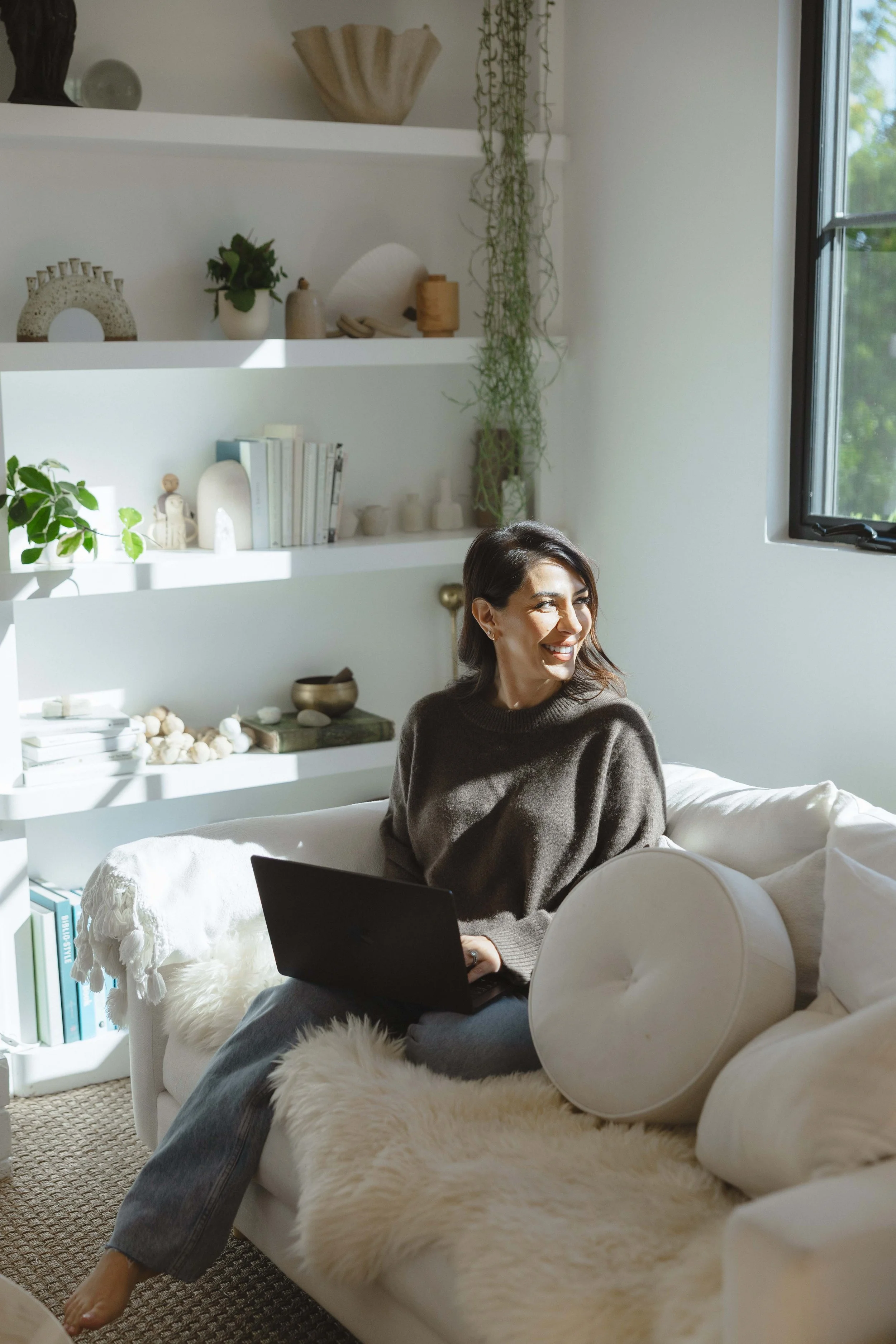 Woman sitting on a beige sofa with a laptop, smiling, in a bright living room with white shelves and window