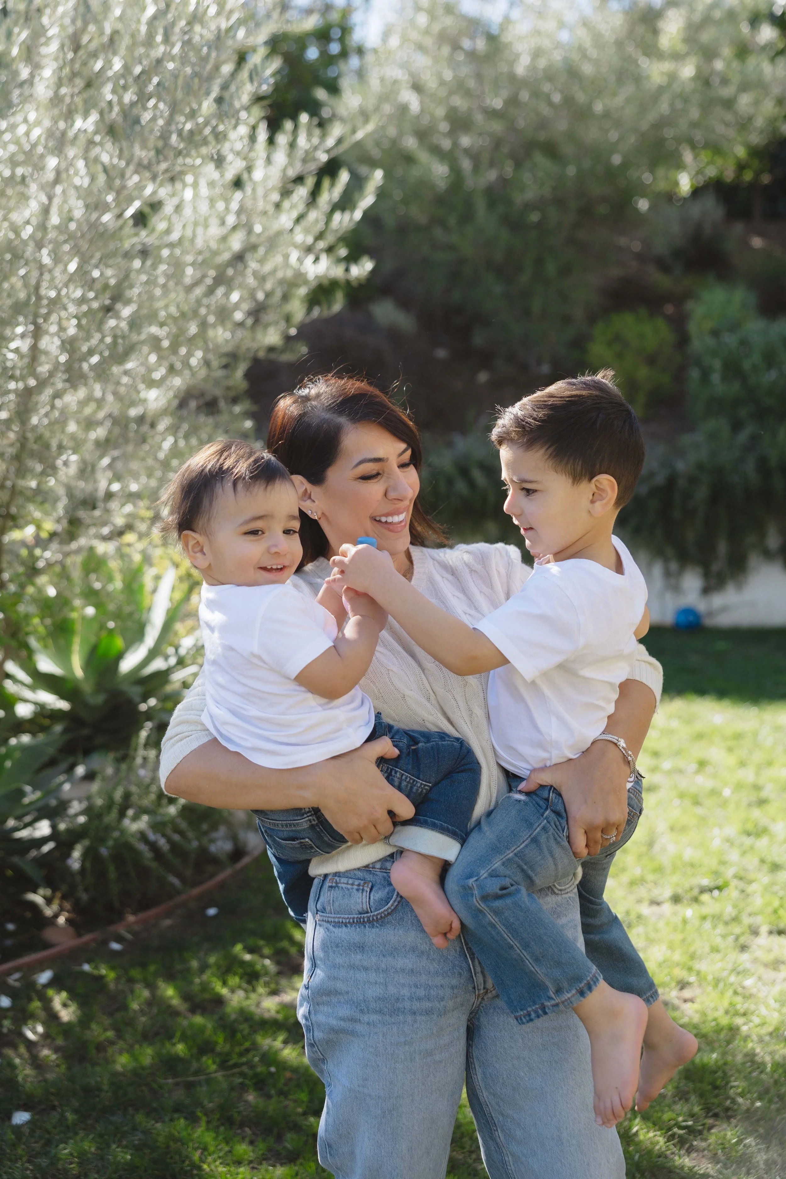 A woman holding two young boys in a garden, smiling and playing during the daytime.