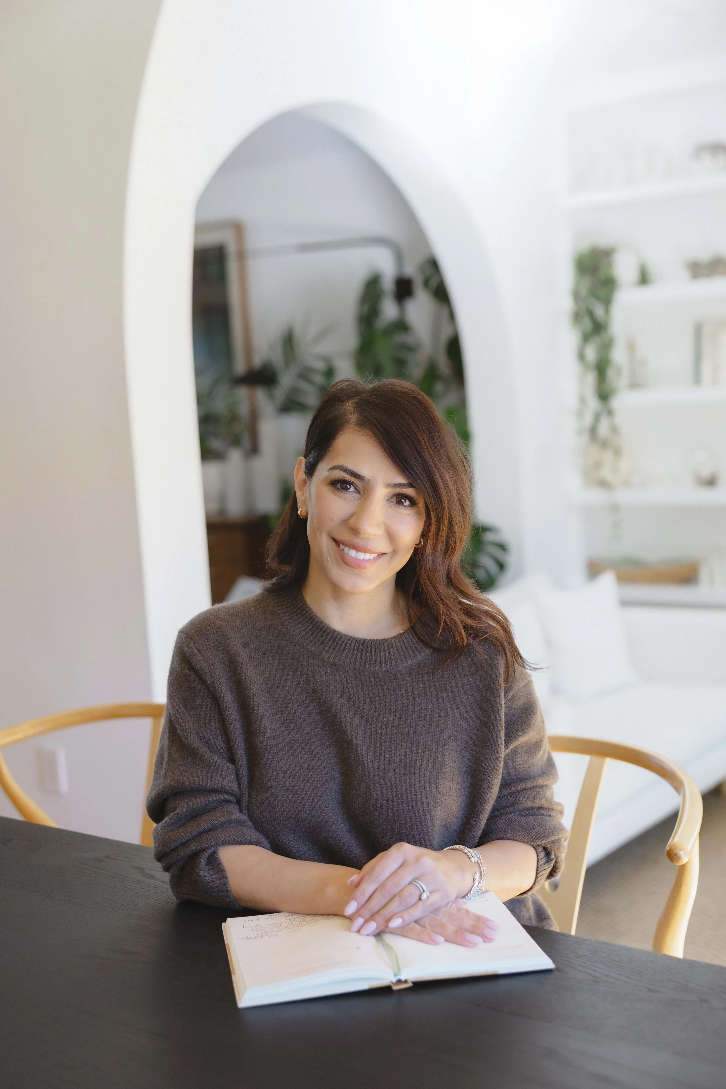 A woman with brown hair, wearing a dark brown sweater, sitting at a dark wooden table with an open notebook in front of her, smiling inside a bright, modern room with white walls, green plants, and shelves.
