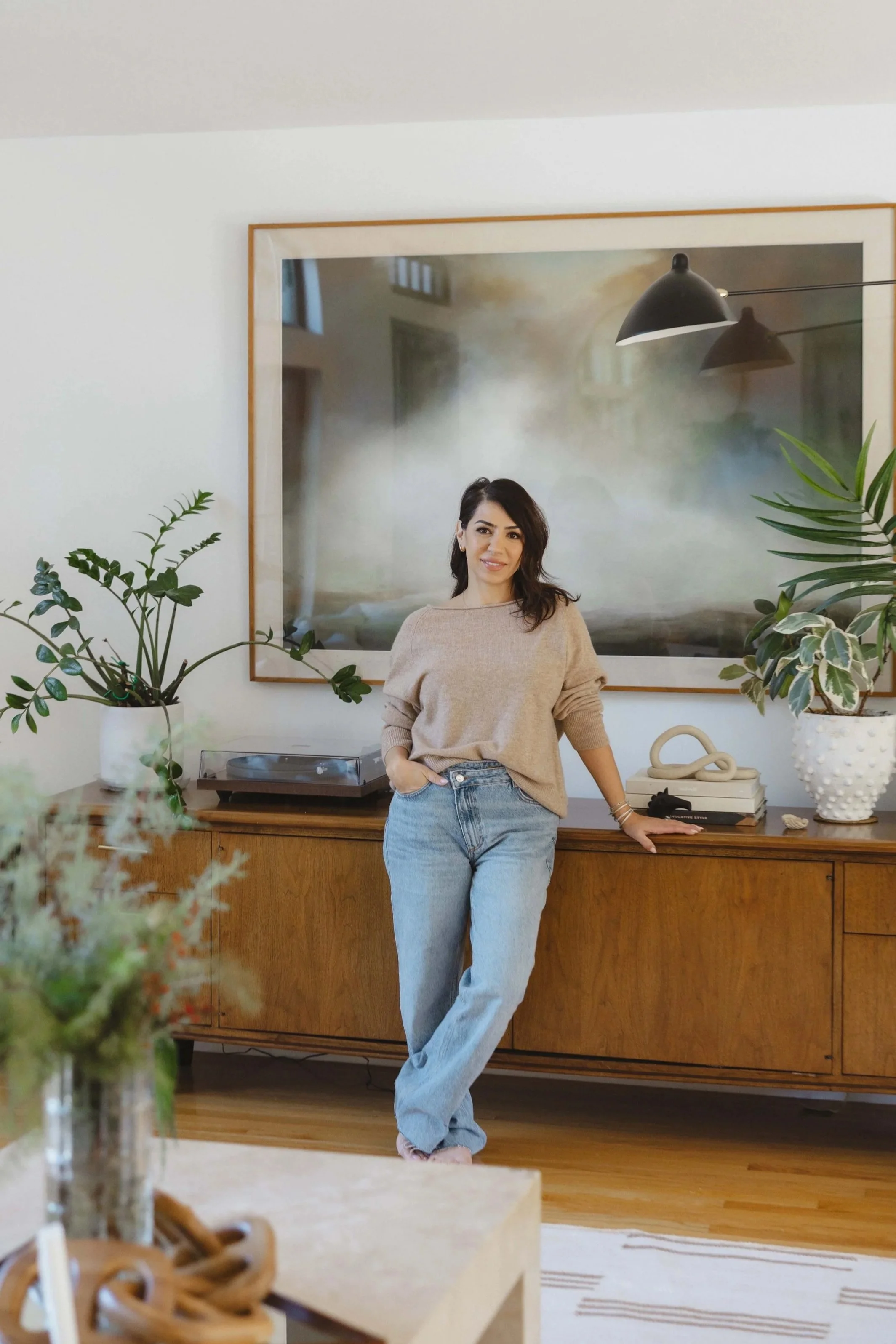 Woman standing in a living room, leaning on a wooden sideboard, with plants and decor, large framed artwork behind her, and wooden flooring.